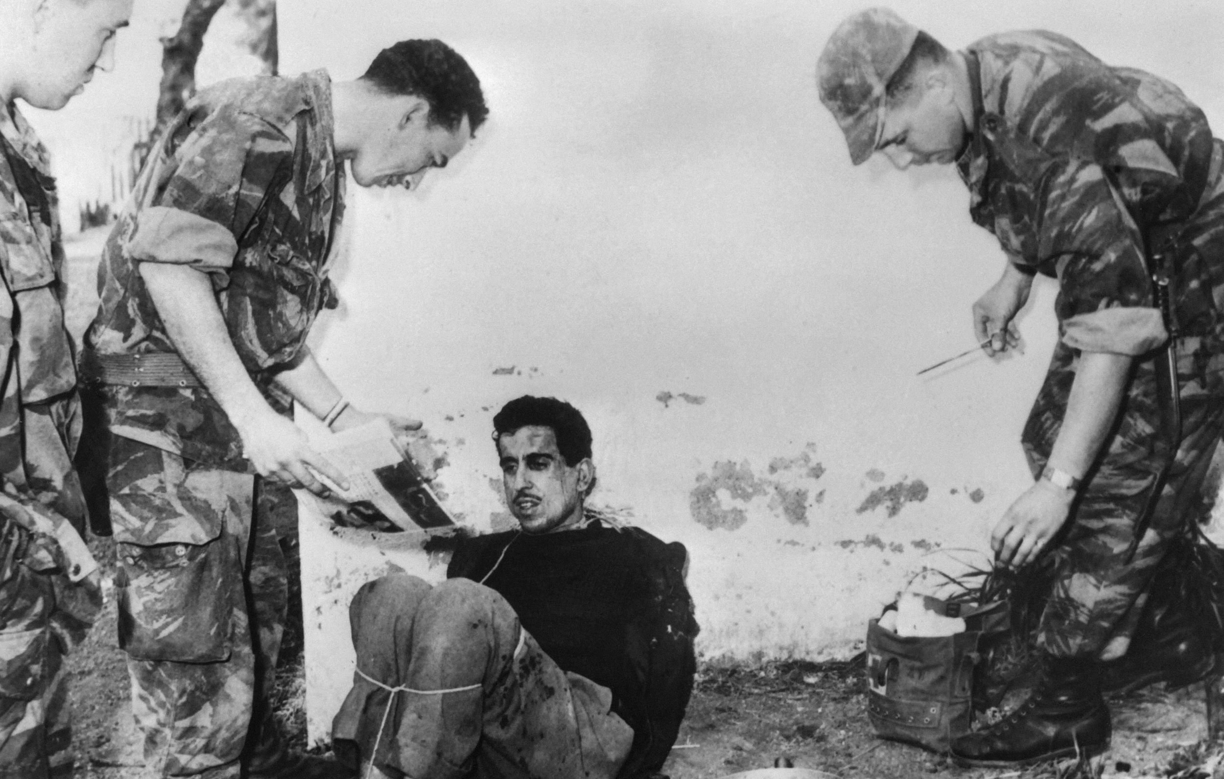 الصورة تظهر مجموعة من الجنود أثناء استجواب رجل يبدو أنه محتجز. الرجل جالس على الأرض ومربوط. الجنود يظهرون بزيهم العسكري، ويبدو أنهم يركزون على الوثائق أو الصور. البيئة المحيطة تبدو خارجية وملوثة بعض الشيء، مما يوحي بمكان غير رسمي. تعكس الصورة أجواء توتر وصعوبة.