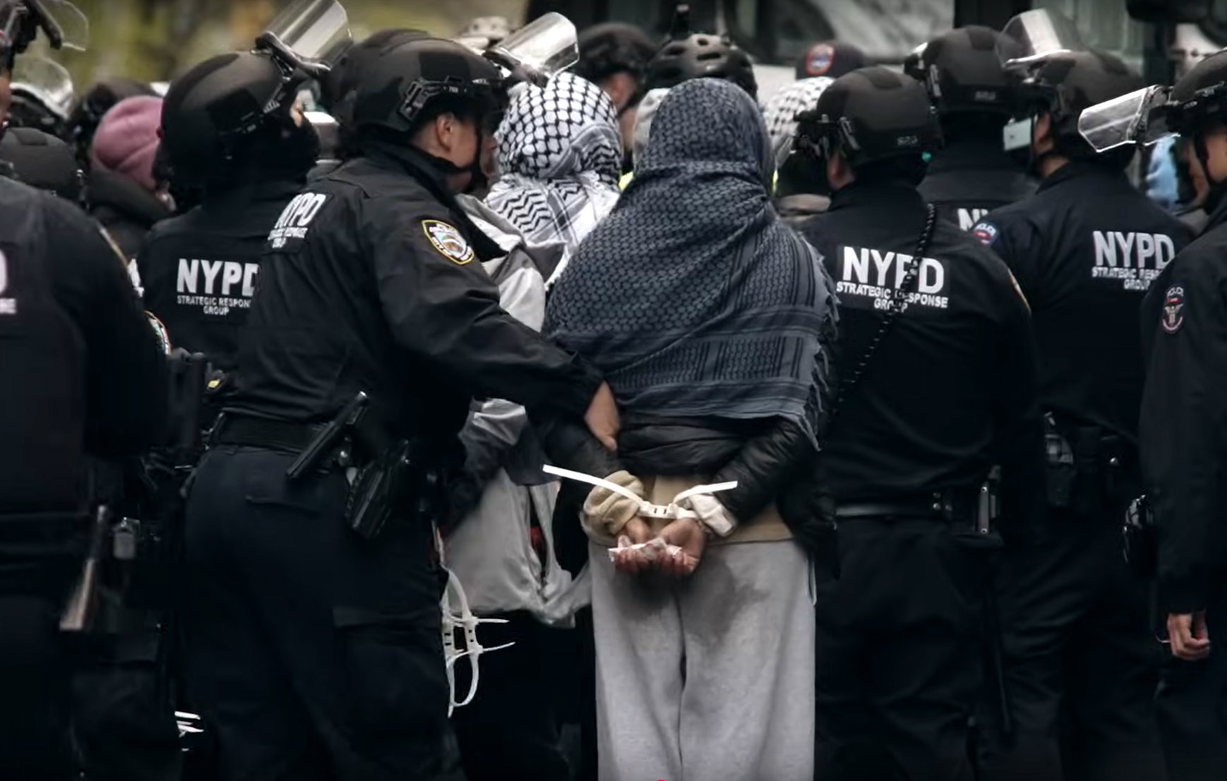 L'image montre un groupe de policiers en uniforme noir entourant une personne arrêtée. Cette personne est de dos, les mains attachées derrière. Les policiers portent des casques et des protections, et il y a une atmosphère tendue, suggérant une manifestation ou une intervention policière.