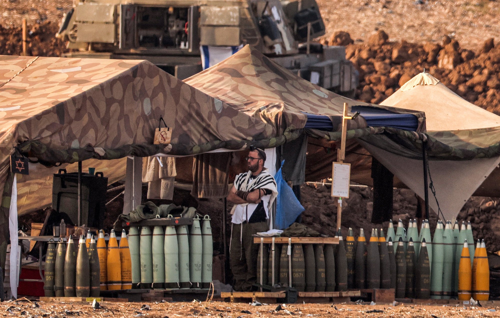 L'image montre un camp militaire en plein air. On y voit des tentes camouflées, et plusieurs obus de différentes tailles sont alignés sur des palettes. Un homme, possiblement en tenue religieuse, se tient debout près des tentes. En arrière-plan, on aperçoit un véhicule militaire. Le sol est poussiéreux, et le paysage semble aride, indiquant une zone de conflit ou d'entraînement militaire.