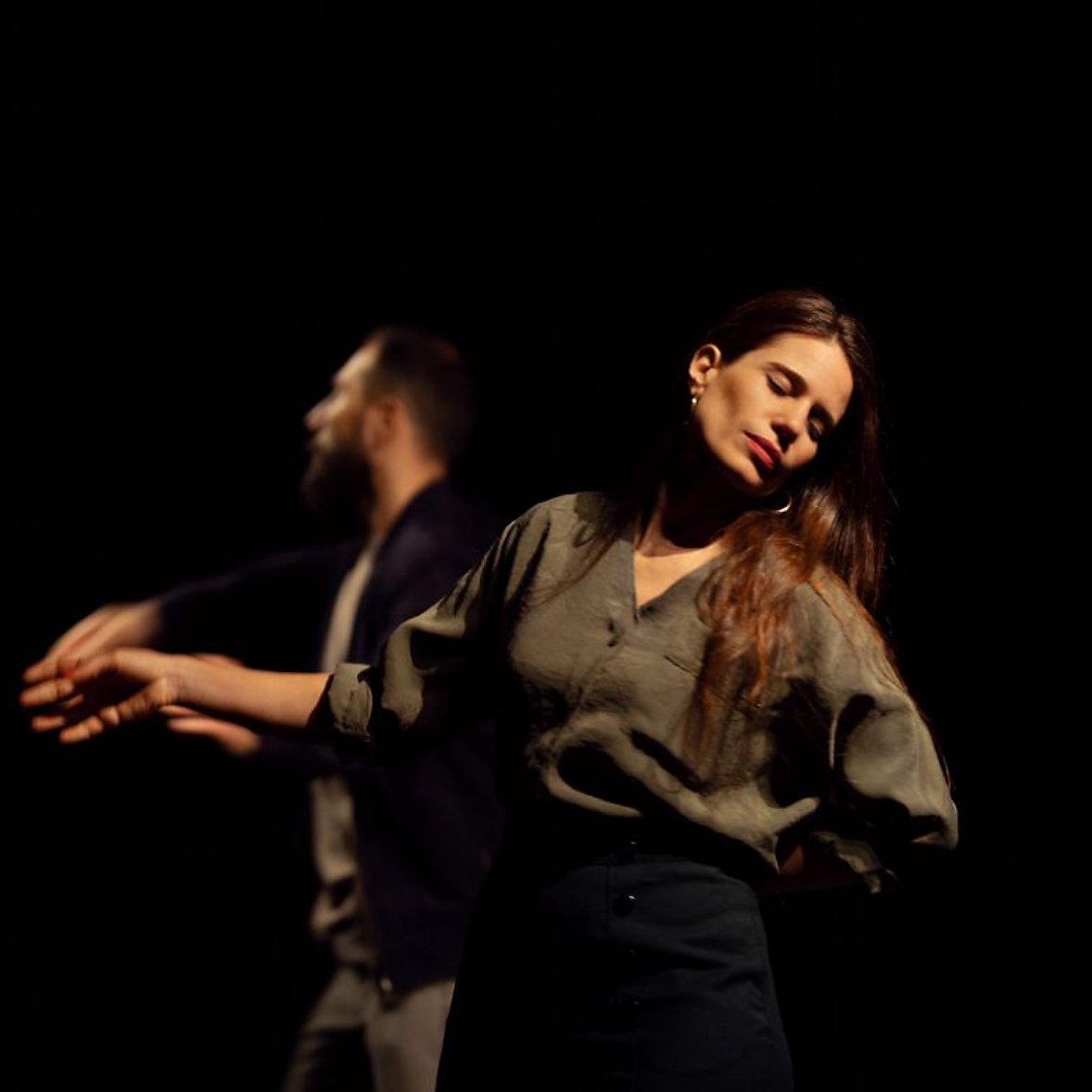 Deux danseurs se déplacent sur scène dans une lumière tamisée. Une femme, les yeux fermés et une expression de sérénité, se tient légèrement en avant, tandis qu'un homme, derrière elle, semble effectuer un mouvement fluide avec ses bras. L'atmosphère est intimiste et expressive.