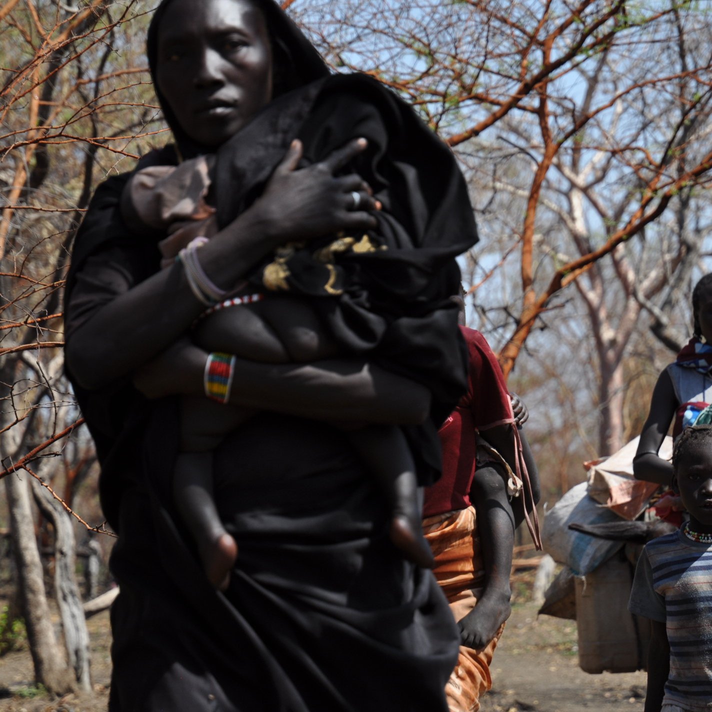 L'image montre un groupe de personnes se déplaçant dans une zone boisée. Au premier plan, une femme porte un enfant sur son dos, tandis qu'à côté d'elle, un petit garçon regarde vers l'avant. D'autres femmes et enfants les suivent, portant des sacs et des objets sur leurs épaules. Leurs vêtements sont colorés, et le décor est constitué d'arbres aux branches dénudées, suggérant un environnement aride. L'atmosphère semble être celle d'un déplacement ou d'une communauté en mouvement.
