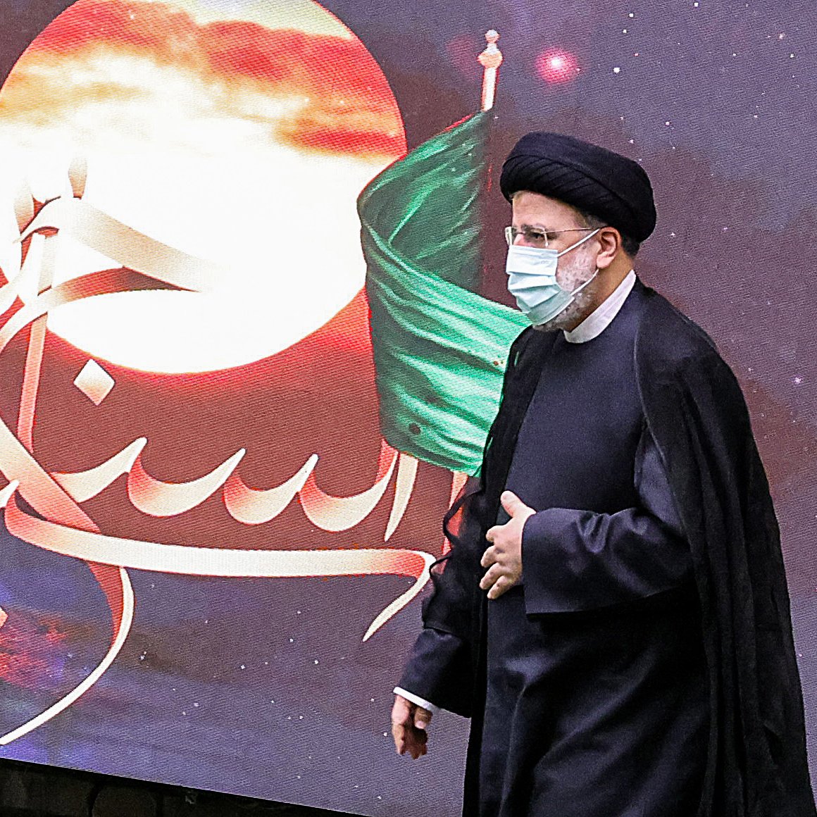 در این تصویر، یک مرد در لباس رسمی و با عمامه مشکی در حال راه رفتن است. او ماسک به صورت دارد و پشت او تصاویری از یک پس‌زمینه هنری با رنگ‌های گرم و نمادهایی مربوط به تاریخ یا فرهنگ دیده می‌شود، از جمله پرچم سبز. متن خوشنویسی شده‌ای در تصویر وجود دارد که به زبان فارسی نوشته شده است. این تصویر ممکن است به یک مراسم مذهبی یا ملی مرتبط باشد.