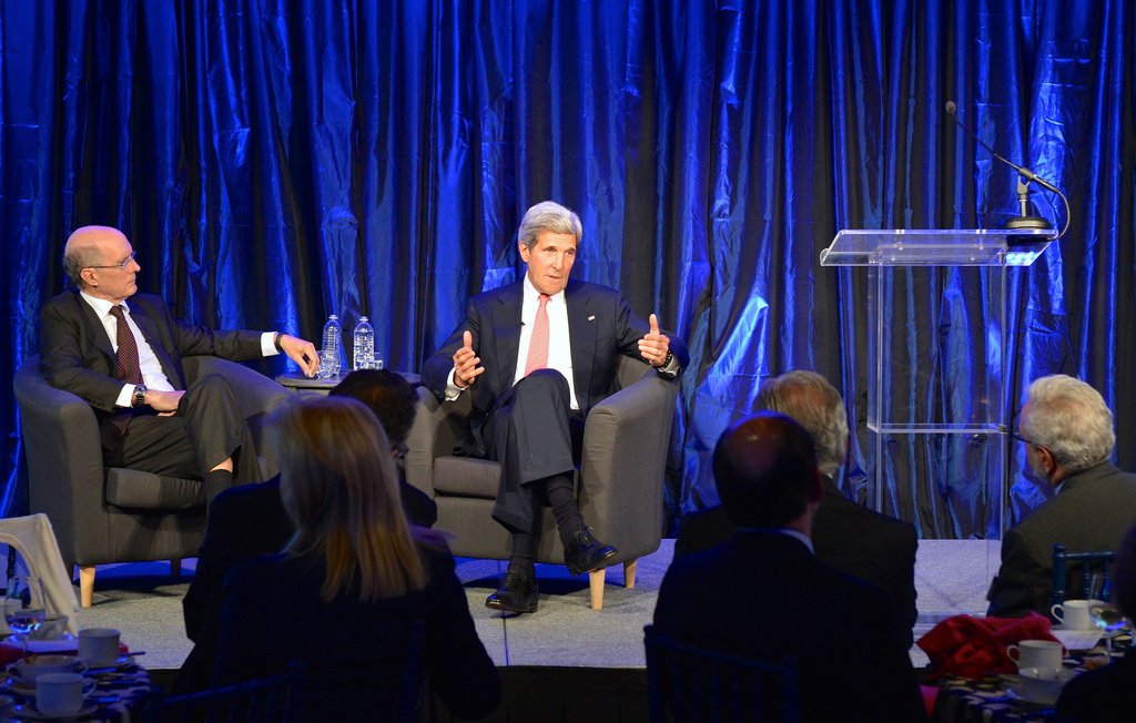 L'image montre une scène de conférence ou de discussion, où deux intervenants sont présents sur une scène. À gauche, un homme est assis sur un fauteuil, écoutant attentivement, tandis qu'à droite, un autre homme parle avec des gestes, probablement en train de partager ses idées ou répondre à des questions. La scène est éclairée avec un fond de rideaux bleus, créant une ambiance professionnelle et formelle. Il y a également un podium avec un microphone, prêt à être utilisé. En bas, on peut apercevoir un public assis, attentif à la conversation.