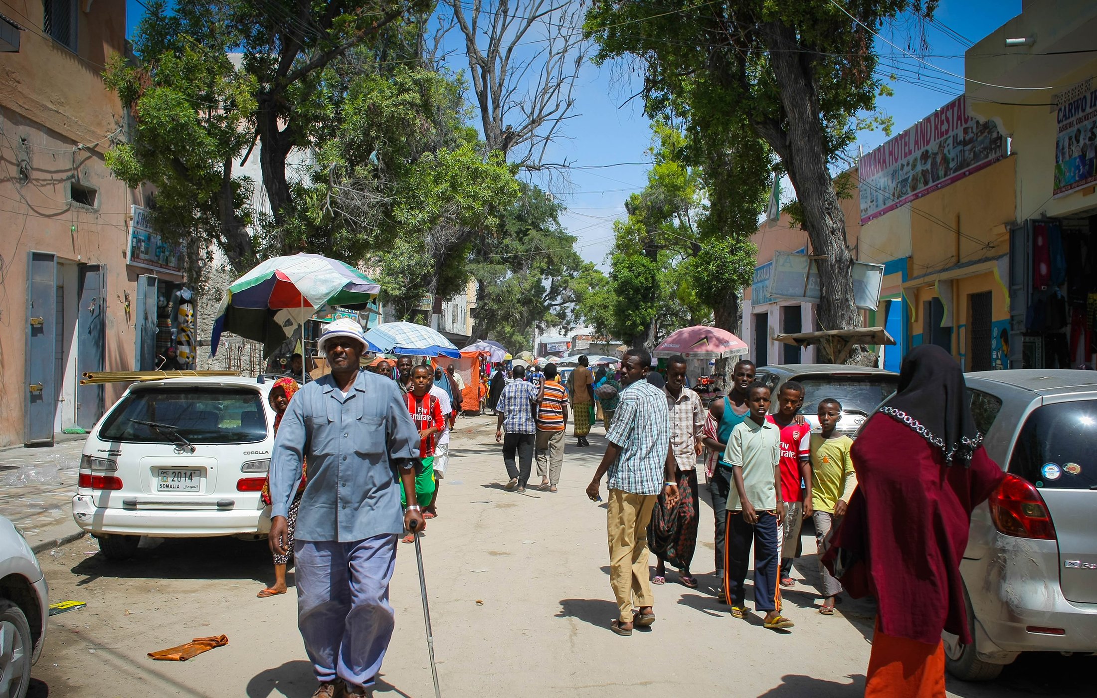 تظهر الصورة شارعا في منطقة حيوية، حيث يسير مجموعة من الأشخاص في اتجاهات مختلفة. في المقدمة، يوجد رجل مسن يستخدم عكازا للمشي، بينما يتواجد أشخاص آخرون في الخلفية، بعضهم يتحدث والبعض الآخر يسير. هناك سيارات متوقفة على الجانبين، وتظلل الأشجار والشمس المكان. يبدو أن الجو مشمس والناس يقومون بأعمالهم اليومية.
