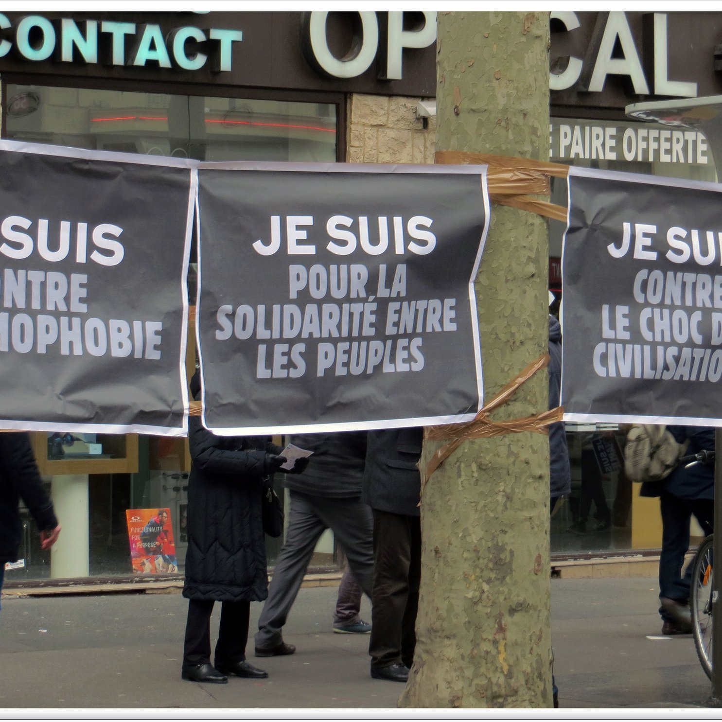 L'image montre plusieurs grandes affiches suspendues, avec des messages écrits en lettres blanches sur fond noir. Les slogans affichés expriment des positions contre l'islamophobie et en faveur de la solidarité entre les peuples. En arrière-plan, on peut apercevoir des personnes qui semblent participer à un rassemblement ou une manifestation. L'ambiance suggère une mobilisation citoyenne pour des causes sociales et politiques.