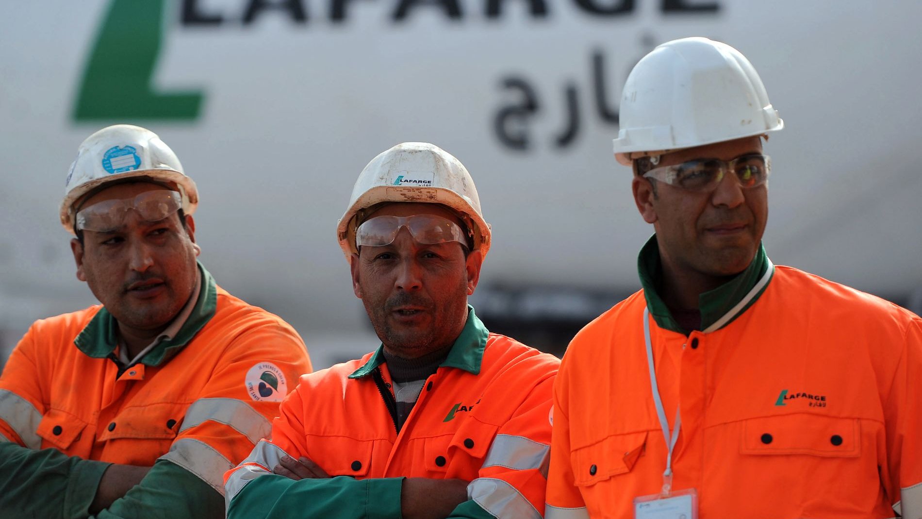 تظهر الصورة ثلاثة رجال يرتدون ملابس العمل البرتقالية، ويضعون خوذات بيضاء على رؤوسهم. يبدو أنهم يعملون في موقع بناء أو مصنع، حيث خلفهم تظهر شاحنة أو معدات تحمل اسم "لافارج". يعبر تعبيرهم عن الجدية والتركيز في العمل، بينما يقفون معًا.