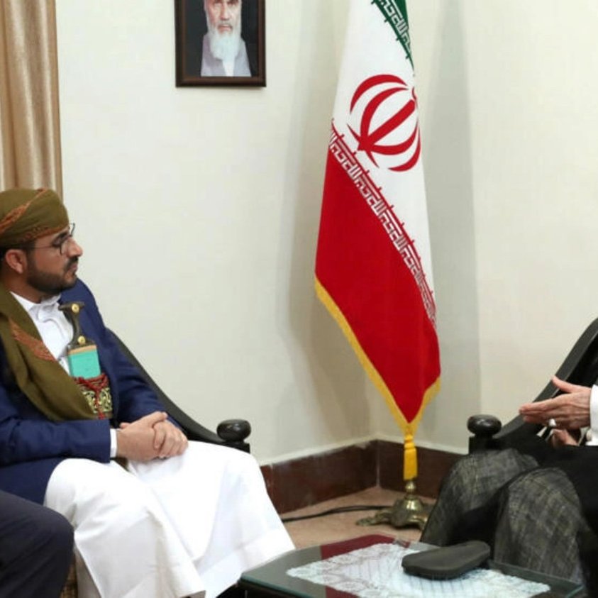 L'image montre une rencontre entre plusieurs personnes dans un bureau. Deux hommes sont assis en face d'un troisième homme qui semble être une personnalité importante, probablement un dirigeant. Les participants portent des vêtements formels et traditionnels. En arrière-plan, on peut voir un drapeau iranien et des rideaux. L'atmosphère semble sérieuse, suggérant un échange diplomatique ou une discussion importante.