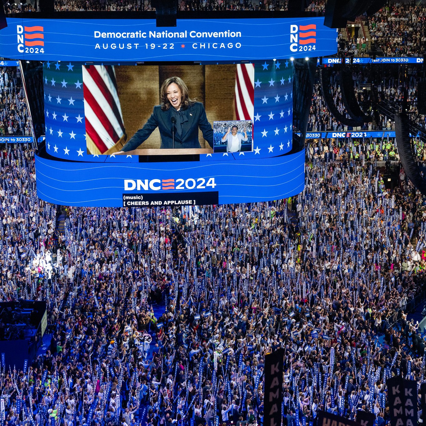 L'image montre une grande scène durant la Convention Nationale Démocrate, qui se déroule à Chicago du 19 au 22 août 2024. On peut voir une foule nombreuse réunie dans une salle, avec des drapeaux et des pancartes en soutien aux candidats. Au centre, un écran géant affiche une intervenante, probablement une personnalité politique, qui s'adresse à l'assemblée. L'atmosphère semble électrisante, avec beaucoup d'enthousiasme parmi les participants.