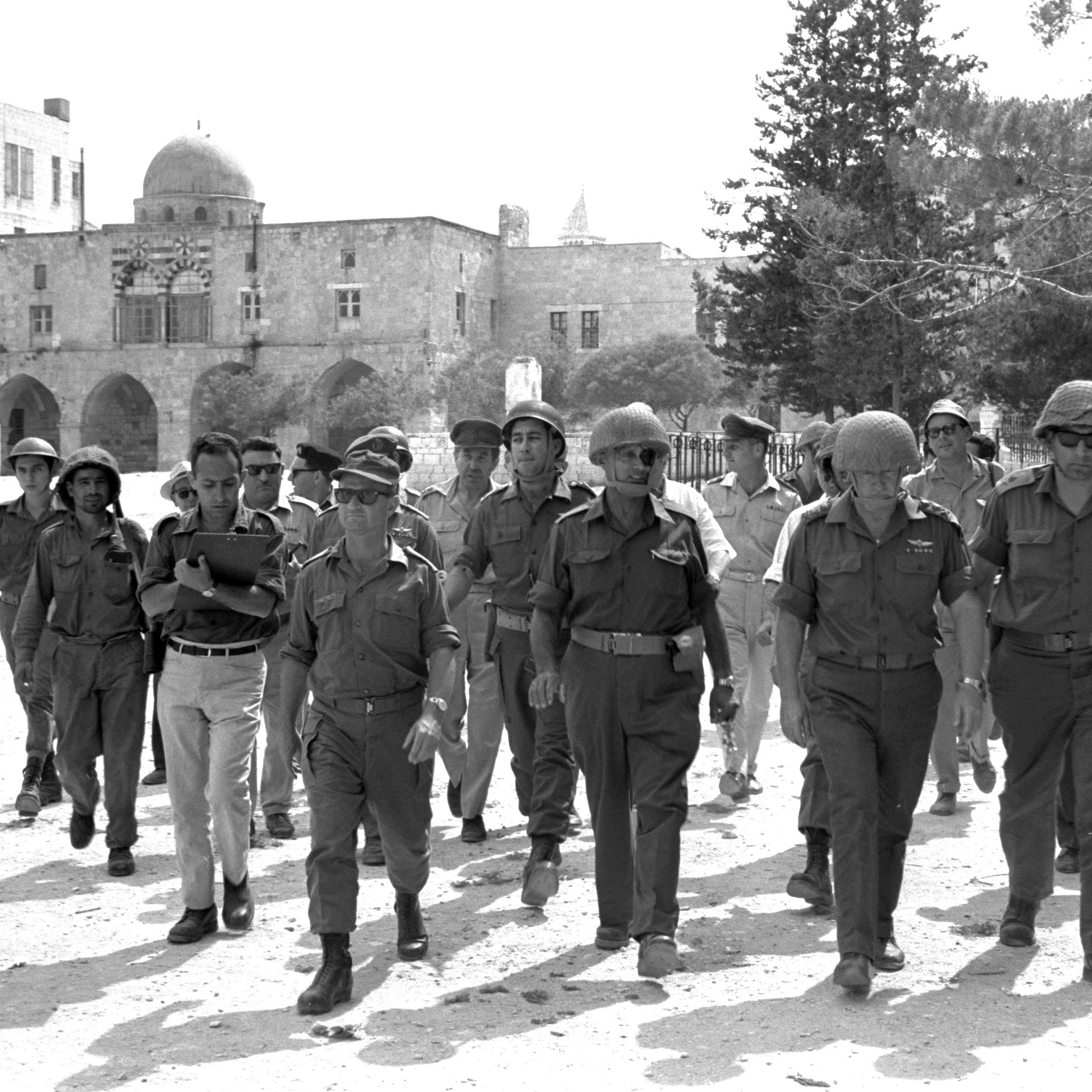 L'image montre un groupe de militaires marchant ensemble dans un environnement urbain. Ils portent des uniformes militaires et des casques. En arrière-plan, on peut voir des bâtiments avec une architecture typique, possiblement historique, ainsi qu'un dôme. L'atmosphère semble sérieuse, avec plusieurs soldats ayant une posture confiante. L'éclairage est lumineux, suggérant qu'il fait soleil. La scène donne une impression de mouvement et de coordination parmi les militaires.