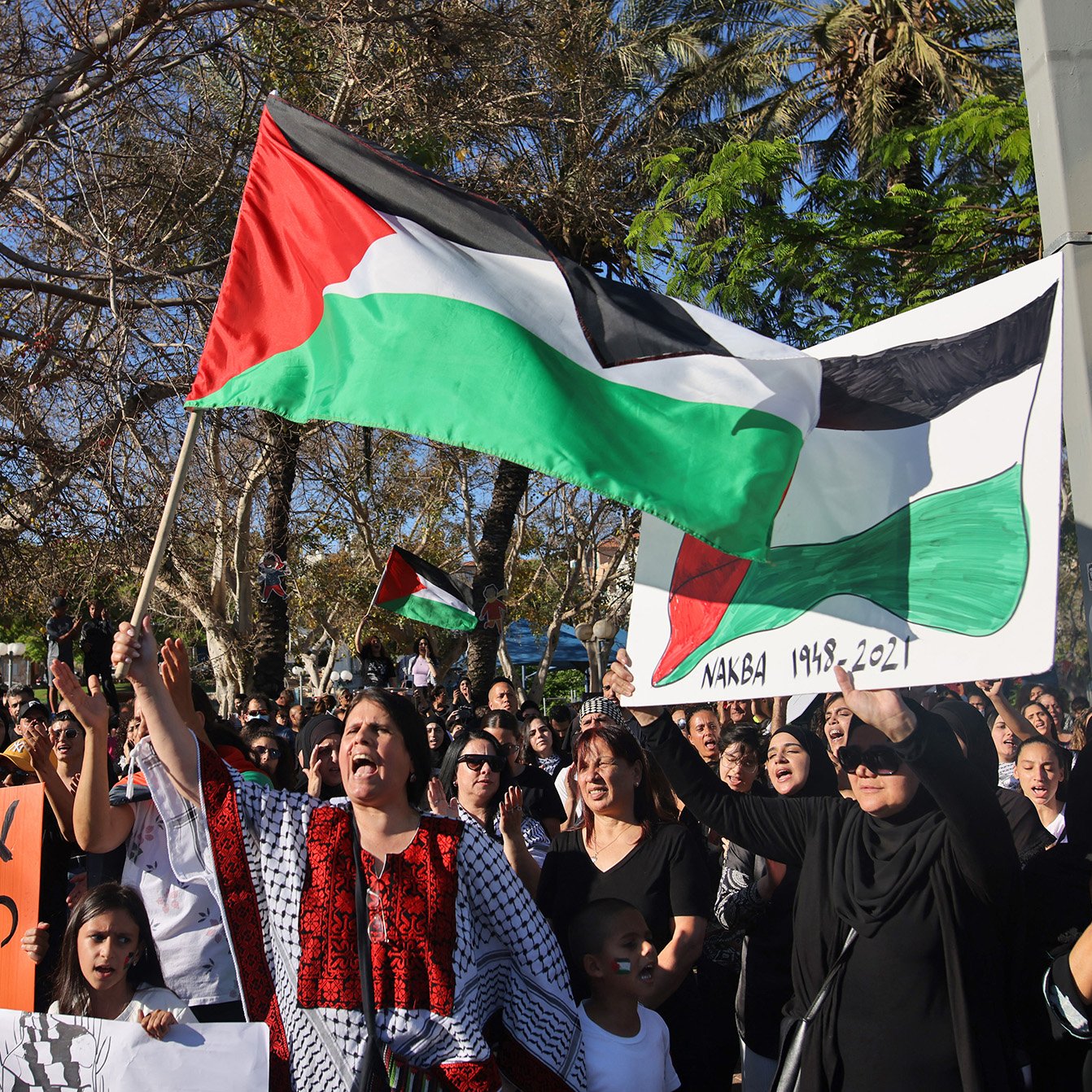 L'image montre une manifestation où des personnes tiennent des drapeaux palestiniens et des pancartes. La foule semble rassemblée pour exprimer une opinion ou un soutien, avec des femmes et des enfants présents. L'arrière-plan inclut des arbres et un environnement urbain. Les participants portent des vêtements variés, certains ayant des motifs traditionnels. L'atmosphère paraît chargée d'émotion et de solidarité.