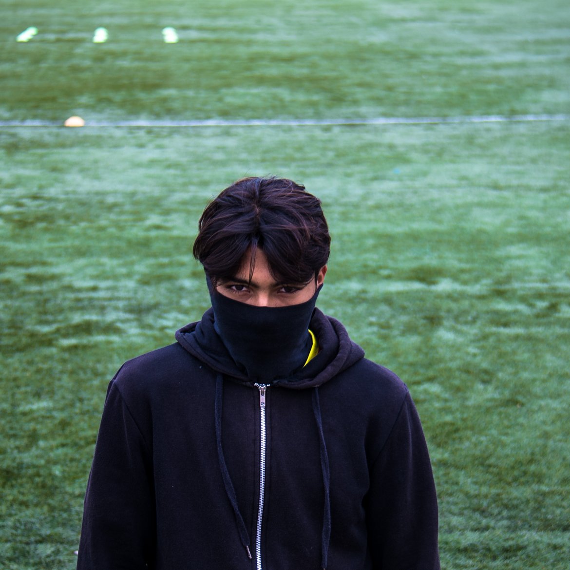 L'image montre un jeune homme se tenant sur un terrain de sport couvert d'herbe. Il porte un sweat à capuche noir et un masque noir qui couvre une partie de son visage. Son regard est intense et fixé vers l'objectif, créant une atmosphère mystérieuse. Le terrain derrière lui est bien entretenu, avec des marques blanches au sol.