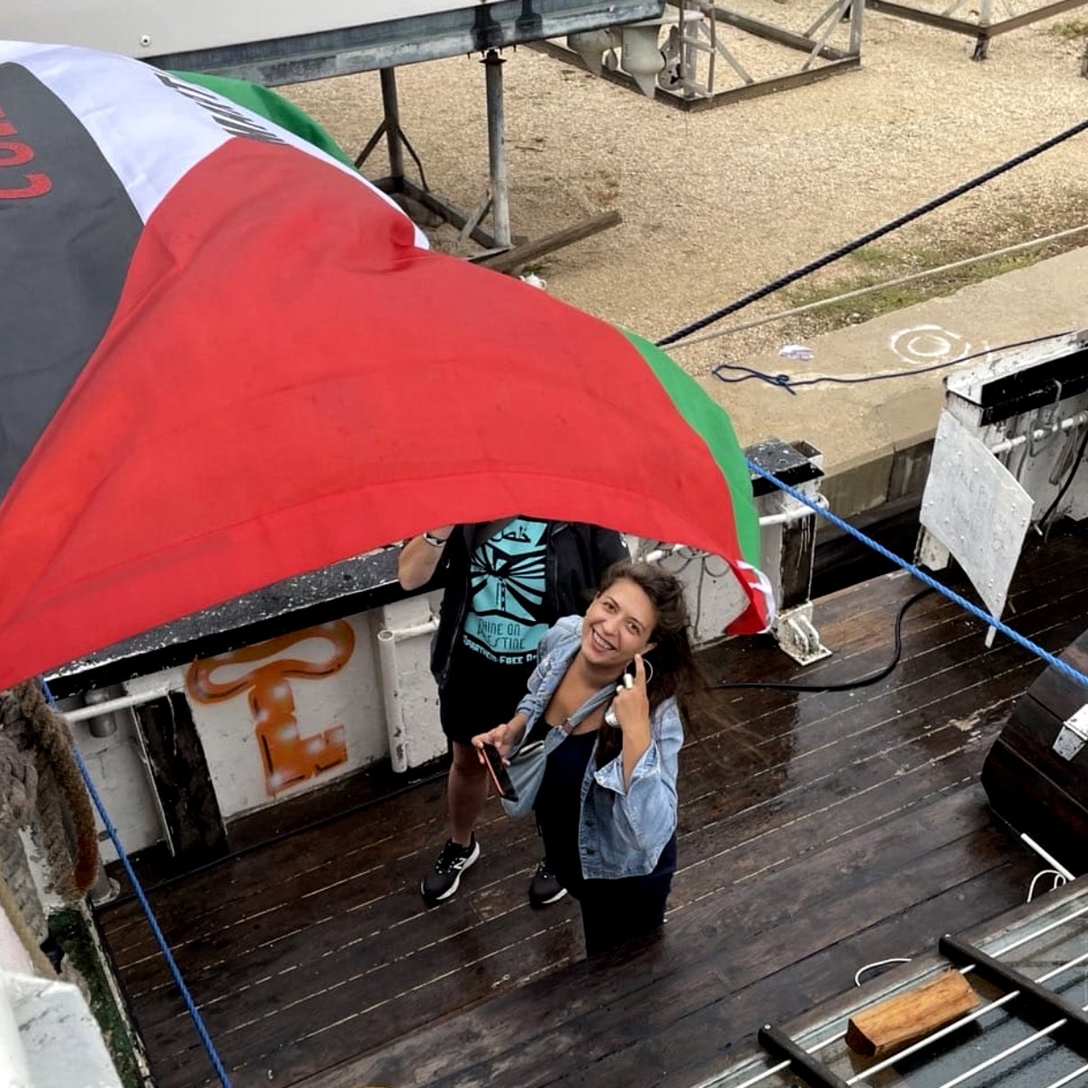 L'image montre deux personnes sur un bateau, sous un grand drapeau noir, rouge et vert. L'une des personnes sourit et pose pour la photo, tandis que l'autre semble tenir le drapeau. Le décor autour inclut du bois, des cordages et d'autres éléments d'un environnement nautique. L'atmosphère semble conviviale et dynamique.