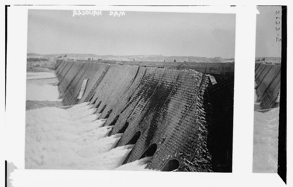 L'image représente un barrage, probablement le barrage d'Assouan, avec une structure en béton. On peut voir de l'eau s'écoulant à travers des ouvertures sur le côté du barrage. Le paysage environnant semble naturel, et le ciel est clair. L'image évoque une impression de puissance hydraulique et d'ingénierie.