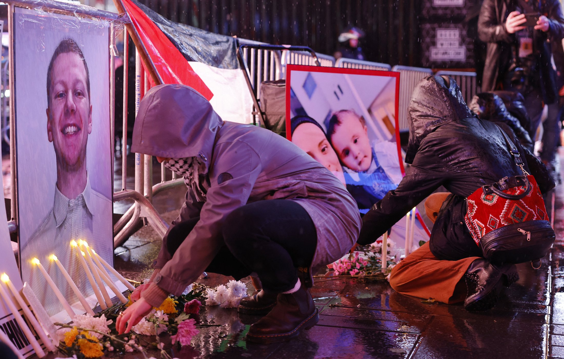 L'image montre des personnes en train de rendre hommage devant des portraits posés au sol. On peut voir des bougies allumées et des fleurs disposées autour des photos. Certaines personnes s'agenouillent ou se penchent pour arranger les fleurs. L'environnement semble humide, peut-être en raison de la pluie, et les participants portent des vêtements adaptés aux intempéries, comme des imperméables et des écharpes. L'atmosphère semble solennelle et empreinte de respect.