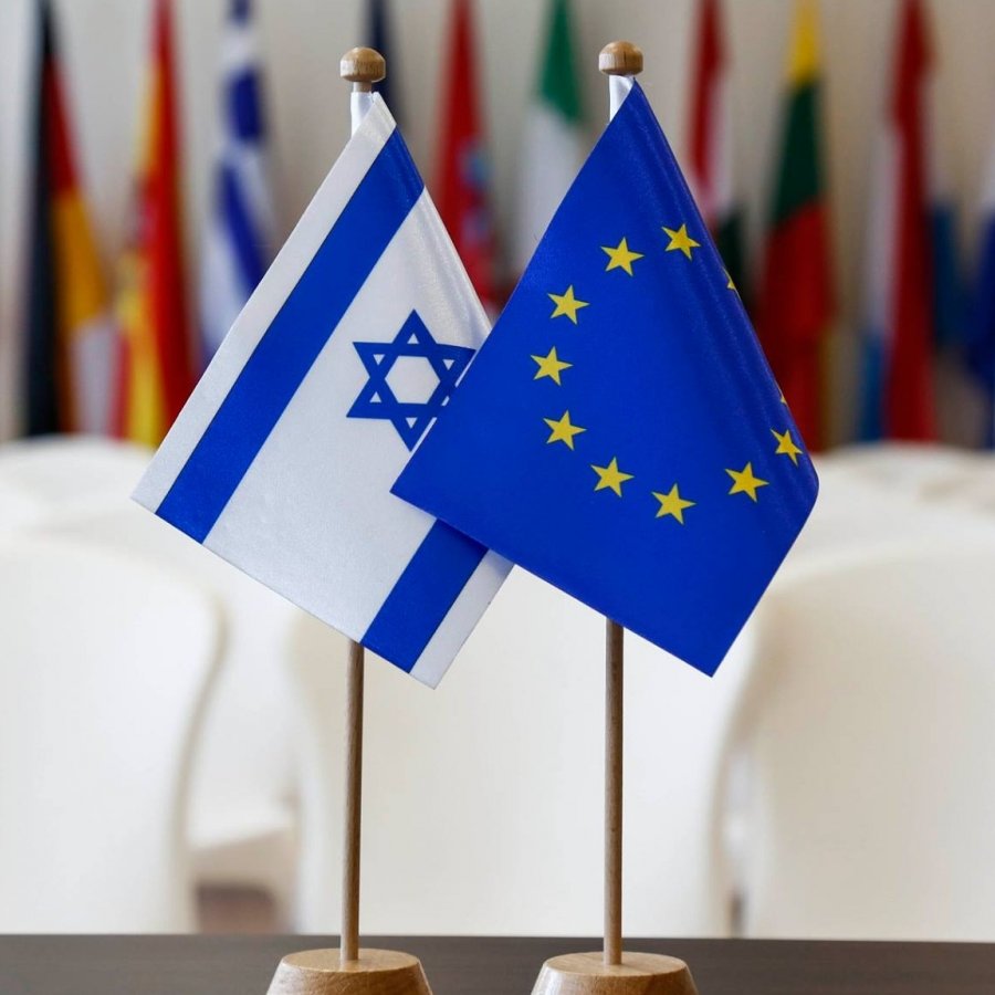 The image features two small flags standing on a table. One flag is the national flag of Israel, which has a blue Star of David and two horizontal blue stripes on a white background. The other flag represents the European Union, displaying a circle of twelve golden stars on a blue background. In the background, there are several other flags, likely from various countries, softly blurred out, suggesting a formal or diplomatic setting.
