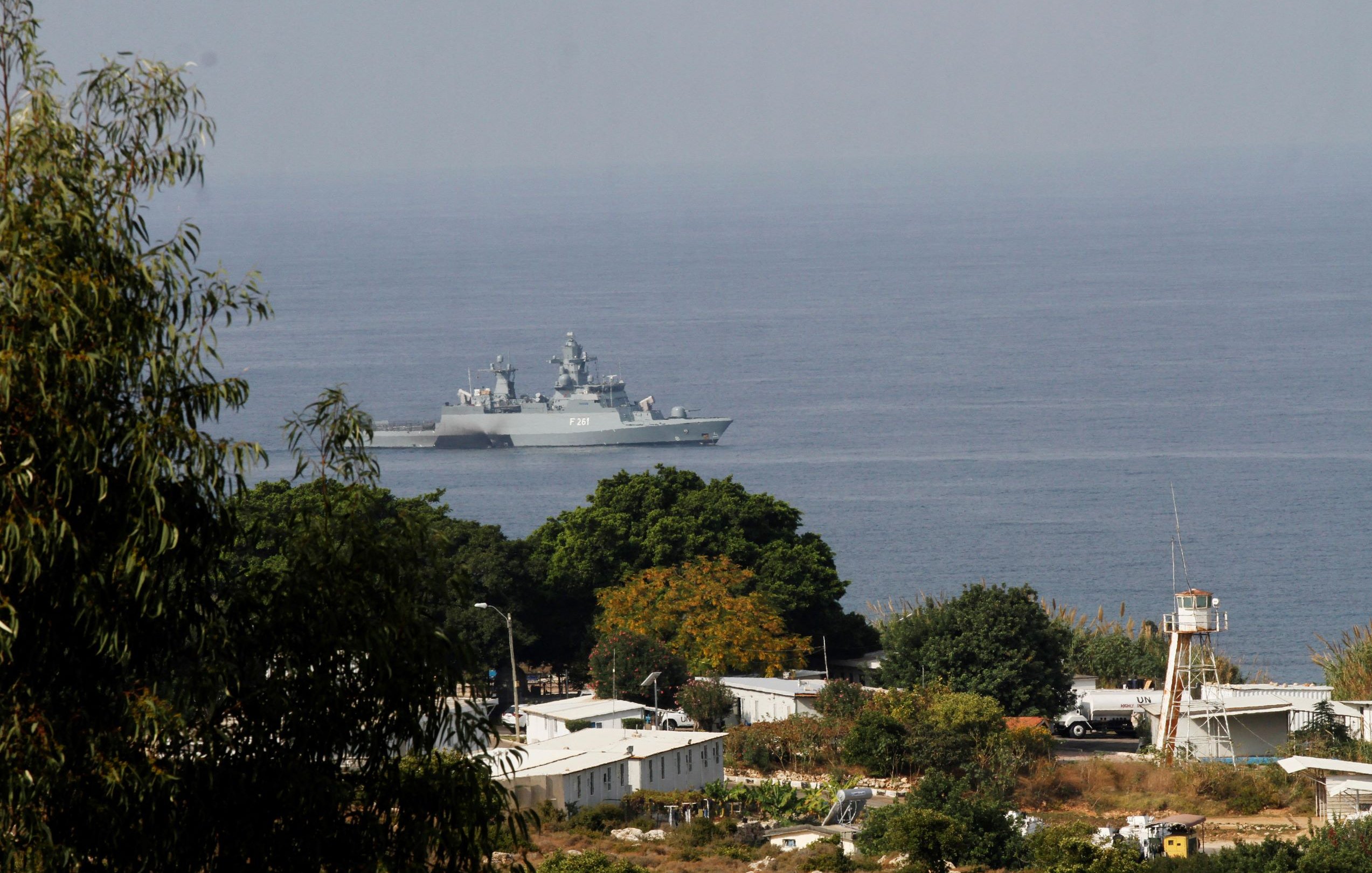 تظهر في الصورة سفينة حربية تجوب المياه بالقرب من ساحل يطل على منطقة خضراء. في الخلفية، يمكن رؤية بعض المباني والمرافق، كما يُلاحظ وجود برج مراقبة. السماء تبدو صافية، والمشهد يعكس أجواء بحرية هادئة.