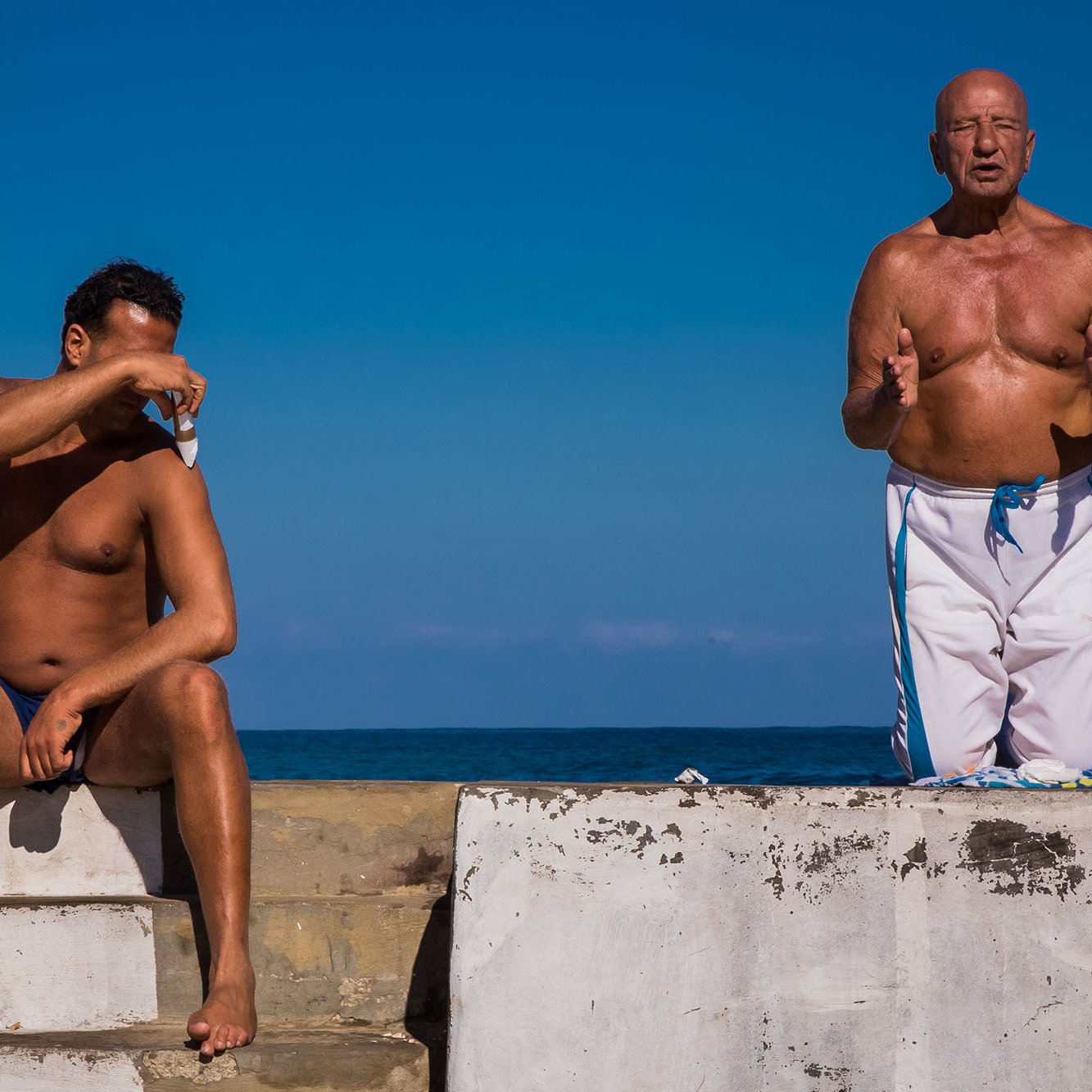 L'image montre deux hommes sur une structure en béton au bord de l'eau. L'un des hommes est assis, torse nu, avec les bras croisés sur un genou, semblant protéger ses yeux du soleil. L'autre homme, debout à côté de lui, est également torse nu et semble parler ou gesticuler, comme s'il était en pleine conversation. En arrière-plan, on peut voir un ciel bleu clair et une étendue de mer, créant une atmosphère estivale et décontractée.