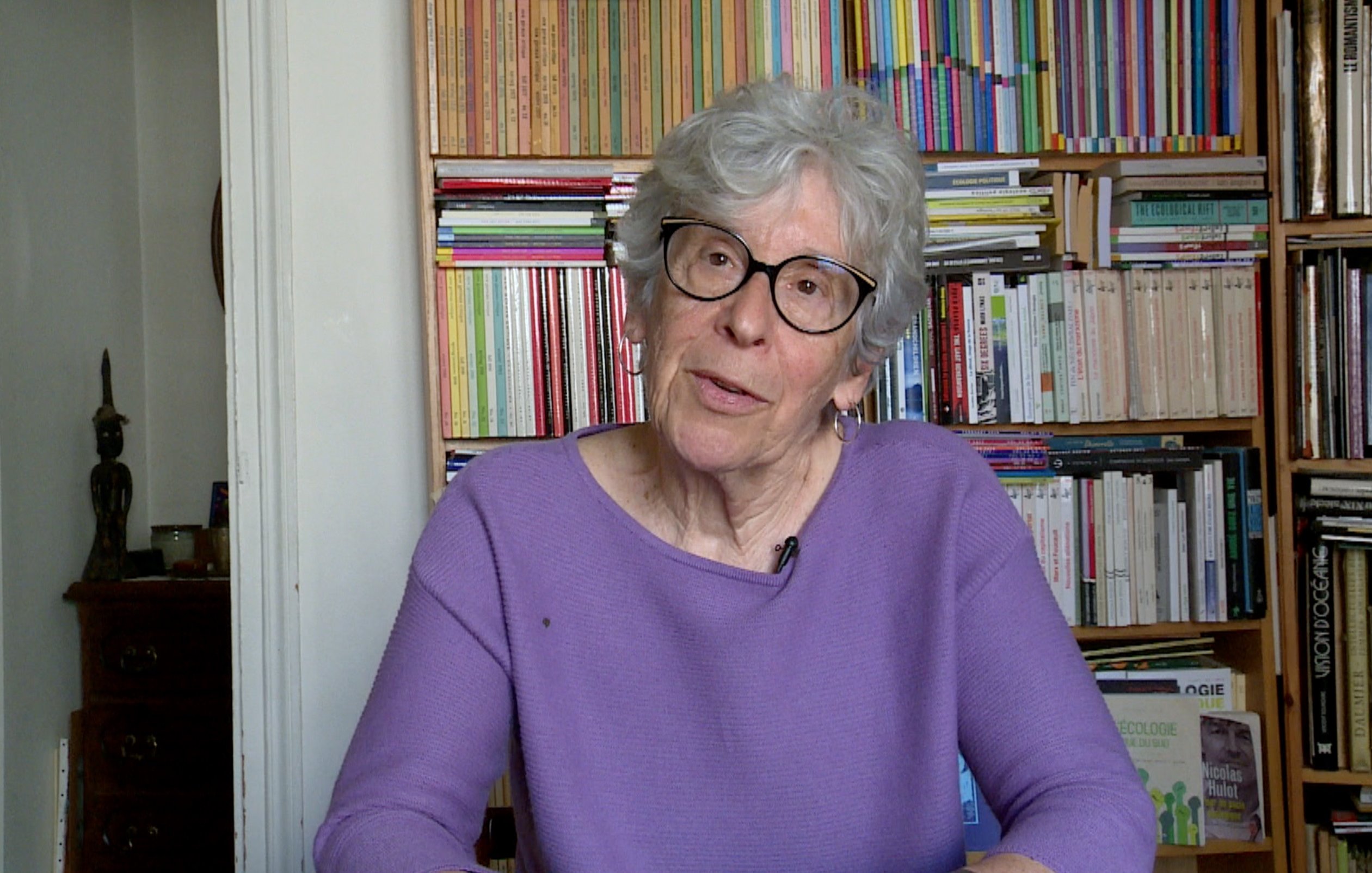 The image features an elderly woman with gray hair and glasses, sitting at a table. She is wearing a purple sweater and appears to be engaged in conversation or sharing her thoughts. In the background, there are bookshelves filled with various books, creating a colorful and organized backdrop. The setting suggests a cozy and intellectual atmosphere.
