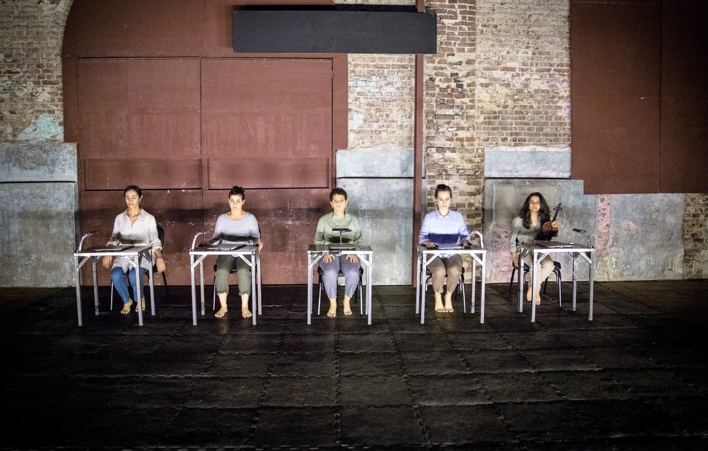 L'image montre un groupe de cinq personnes assises sur des chaises, disposées en ligne. Elles portent des vêtements simples et sont pieds nus, ce qui donne une impression de simplicité et d'intimité. L'environnement est industriel, avec des murs en brique apparente et une lumière tamisée qui crée une ambiance à la fois contemplative et moderne. Les personnes semblent concentrées, chacune avec un objet devant elle, suggérant un moment de performance ou de réflexion partagée.