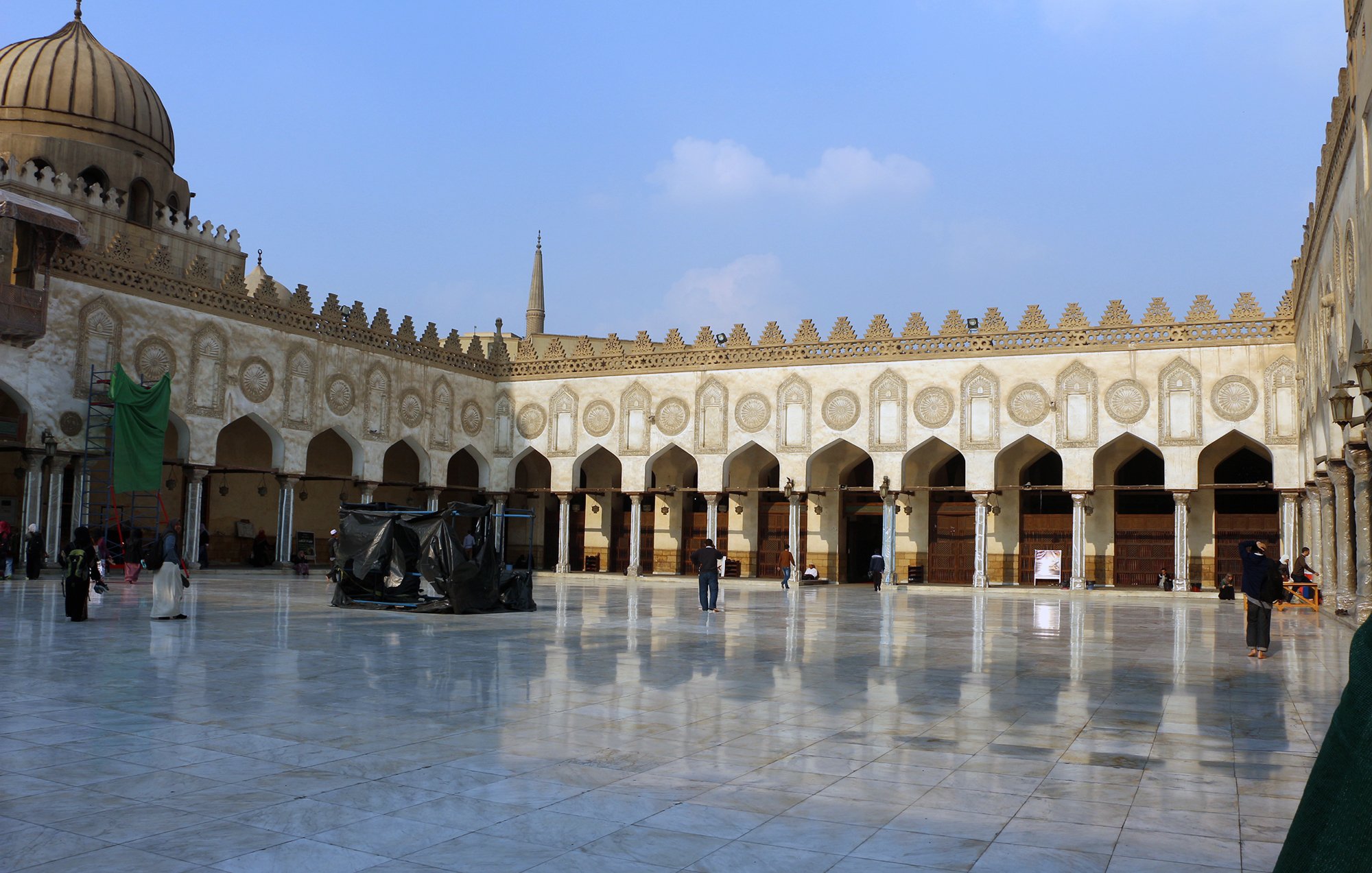 L'image montre une cour spacieuse d'un bâtiment historique, probablement un lieu de culte ou une mosquée. Le sol est en marbre poli, reflétant la lumière du ciel. On peut observer des arches élégantes et de nombreuses colonnes tout autour, ornées de motifs architecturaux détaillés. Au fond, on aperçoit un dôme et des minarets qui s'élèvent dans le ciel. La scène est calme, avec quelques personnes se déplaçant dans l'espace. Le ciel est clair, parsemé de nuages légers.