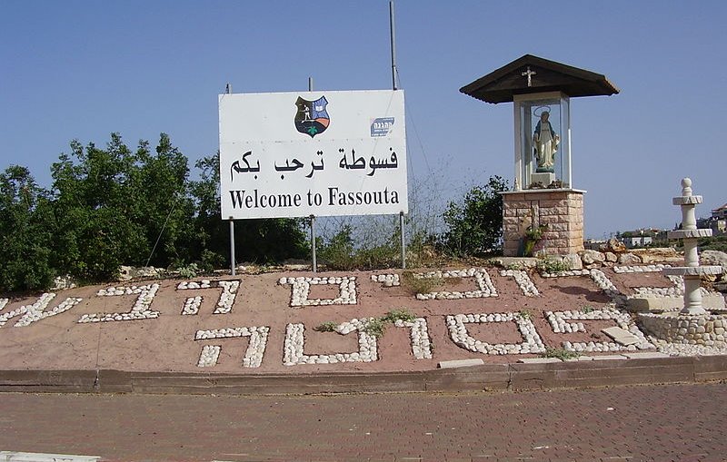 L'image montre un panneau de bienvenue à Fassouta, une localité. Le panneau est écrit en arabe et en anglais. À côté du panneau, il y a un abri qui semble abriter une statue, probablement de la Vierge Marie, indiquant une certaine importance religieuse à cet endroit. Le sol est décoré avec des mots formés de cailloux blancs, ajoutant une touche artisanale à l'ensemble. En arrière-plan, on aperçoit des arbres ou de la verdure, donnant une impression de nature environnante.