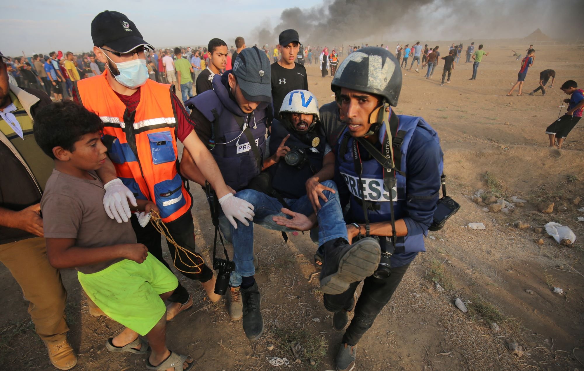 L'image montre une scène dramatique où plusieurs personnes, dont des journalistes, interviennent dans une situation d'urgence. Un homme blessé est transporté par des secouristes, tandis qu'un jeune garçon semble les observer. En arrière-plan, il y a des fumées, probablement d'incendies, et une foule rassemblée. L'environnement semble chargé d'émotion et de tension, évoquant des événements majeurs ou des conflits.