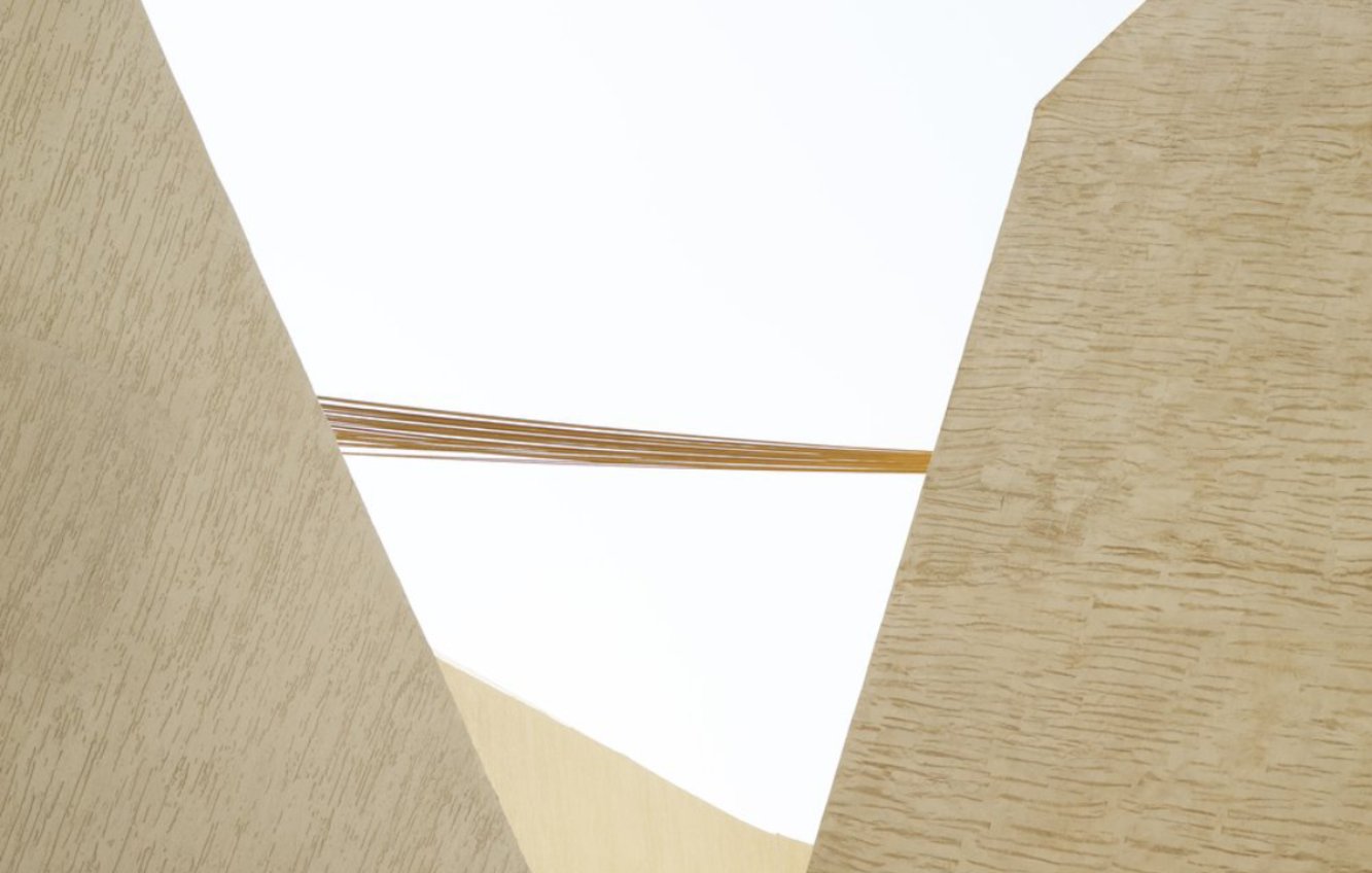 L'image montre une composition architecturale contemporaine, avec des murs en forme de vagues ou de angles vifs. Les surfaces sont d'un ton clair, très neutre, et présentent une texture subtile. Des fils ou des câbles sont tendus au-dessus, créant une interaction visuelle intéressante entre les formes géométriques des murs et les lignes horizontales des câbles. L'ensemble dégage une impression de modernité et de légèreté, tout en jouant sur les contrastes de formes et de matériaux. Le fond est clair, ce qui accentue la luminosité de la scène.