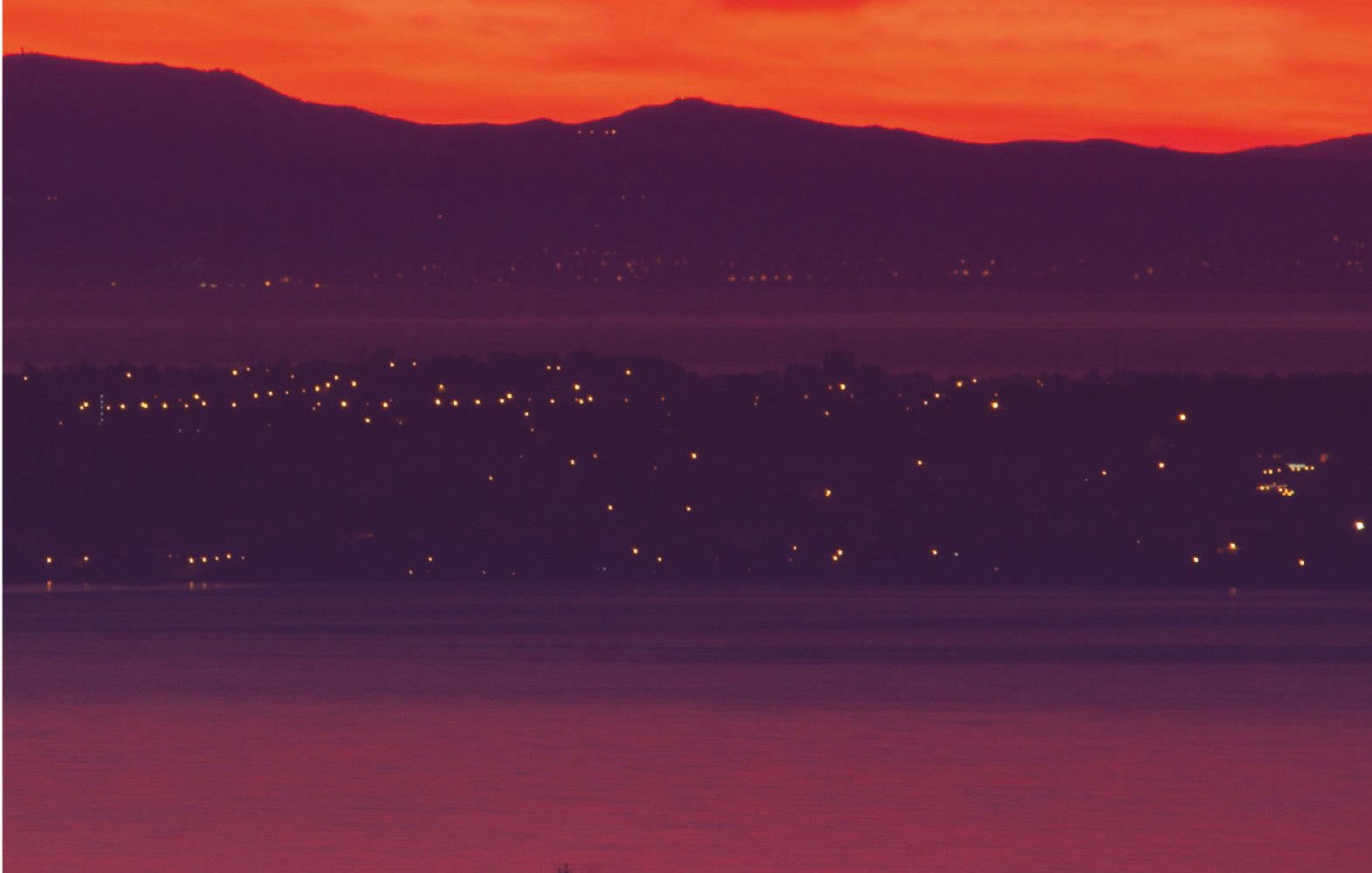 L'image présente un coucher de soleil spectaculaire, où le ciel est teinté de rouge et d'orange lumineux. En arrière-plan, on aperçoit des collines sombres, tandis qu'à l'avant-plan, un cargo navigue sur l'eau calme. Les lumières d'une ville ou d'un port scintillent le long de la côte, ajoutant une touche de vie à cette scène paisible.