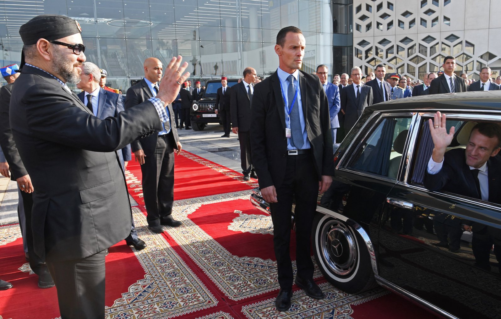 تظهر الصورة مجموعة من الأشخاص في حدث رسمي. يوجد فيها شخصان بارزان، أحدهما يبدو أنه يلوح بيده، بينما الآخر يجلس داخل سيارة ويقوم أيضًا بالتحية. الخلفية تتضمن مبنى عصري، وهناك سجاد ملون على الأرض، مما يدل على أهمية المناسبة. كما يبدو أن هناك حراسة أمنية حول الشخصيات الحاضرة.