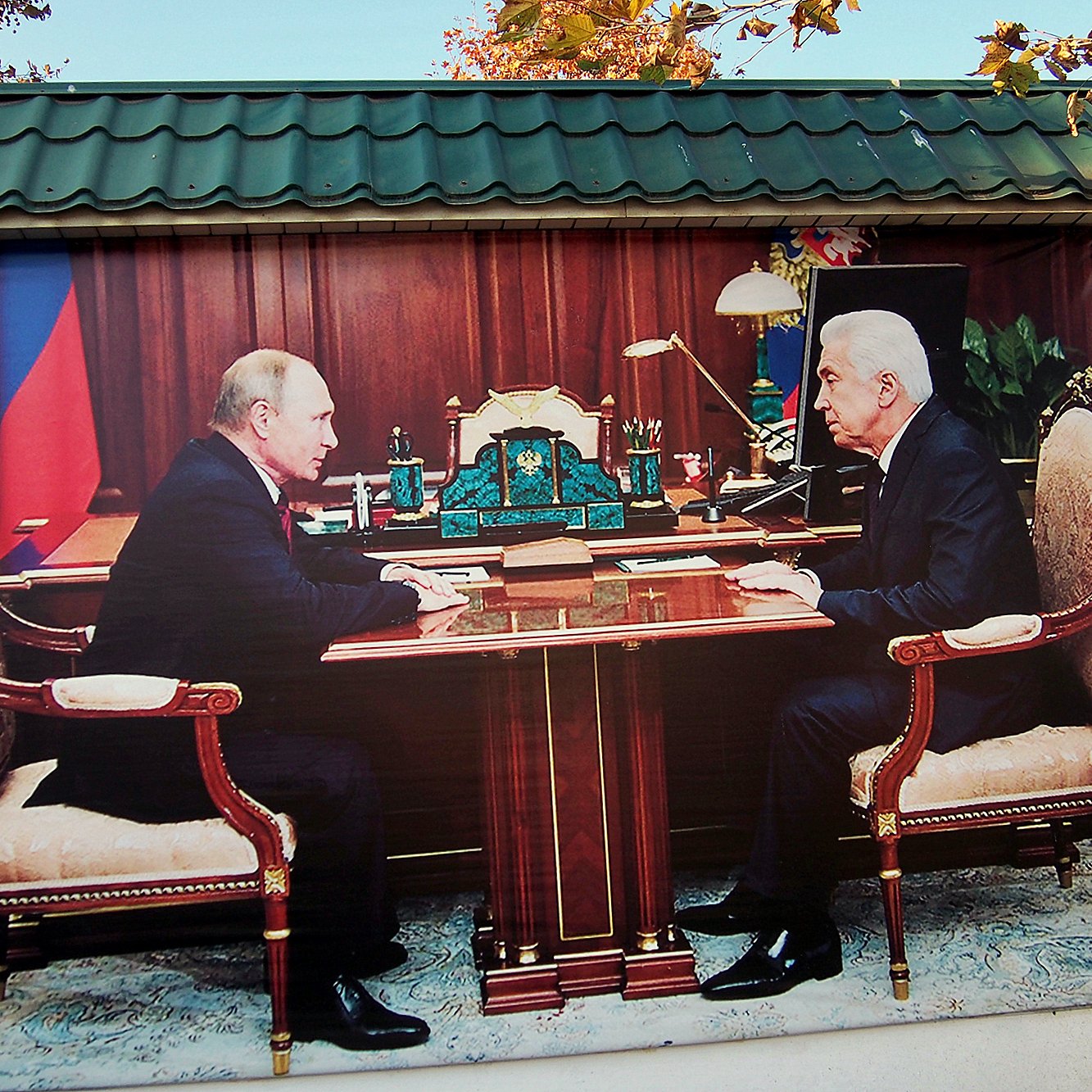 L'image montre deux hommes assis face à face à un bureau dans un bureau luxueux. Ils semblent s'engager dans une conversation sérieuse. Le décor est riche, avec des éléments de mobilier élégants et des accessoires de bureau. En arrière-plan, on peut apercevoir une grande fenêtre qui laisse entrevoir un paysage montagnard.
