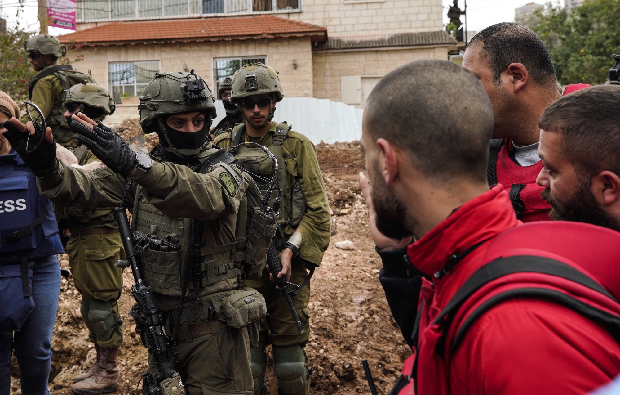 Nell'immagine ci sono diversi uomini in un contesto urbano, alcune persone indossano giacche protettive e una di loro è in uniforme militare con un'arma. Sembra esserci una discussione in corso tra le persone. Il terreno attorno a loro è fangoso e c'è un edificio sullo sfondo.