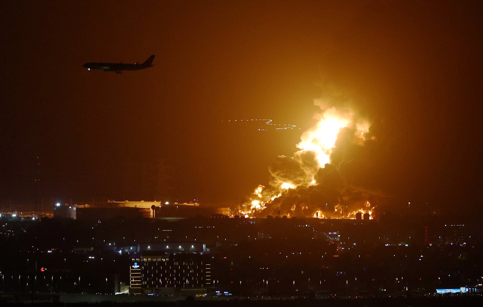 The image depicts a dramatic nighttime scene featuring a large, fiery blaze, likely arising from an industrial area. Bright flames and thick smoke illuminate the dark sky, creating a stark contrast against the surrounding darkness. In the foreground, there are city lights visible, indicating urban development, while an airplane can be seen flying overhead, possibly approaching a nearby airport. The scene suggests a serious incident, such as an explosion or fire, with significant visual impact.