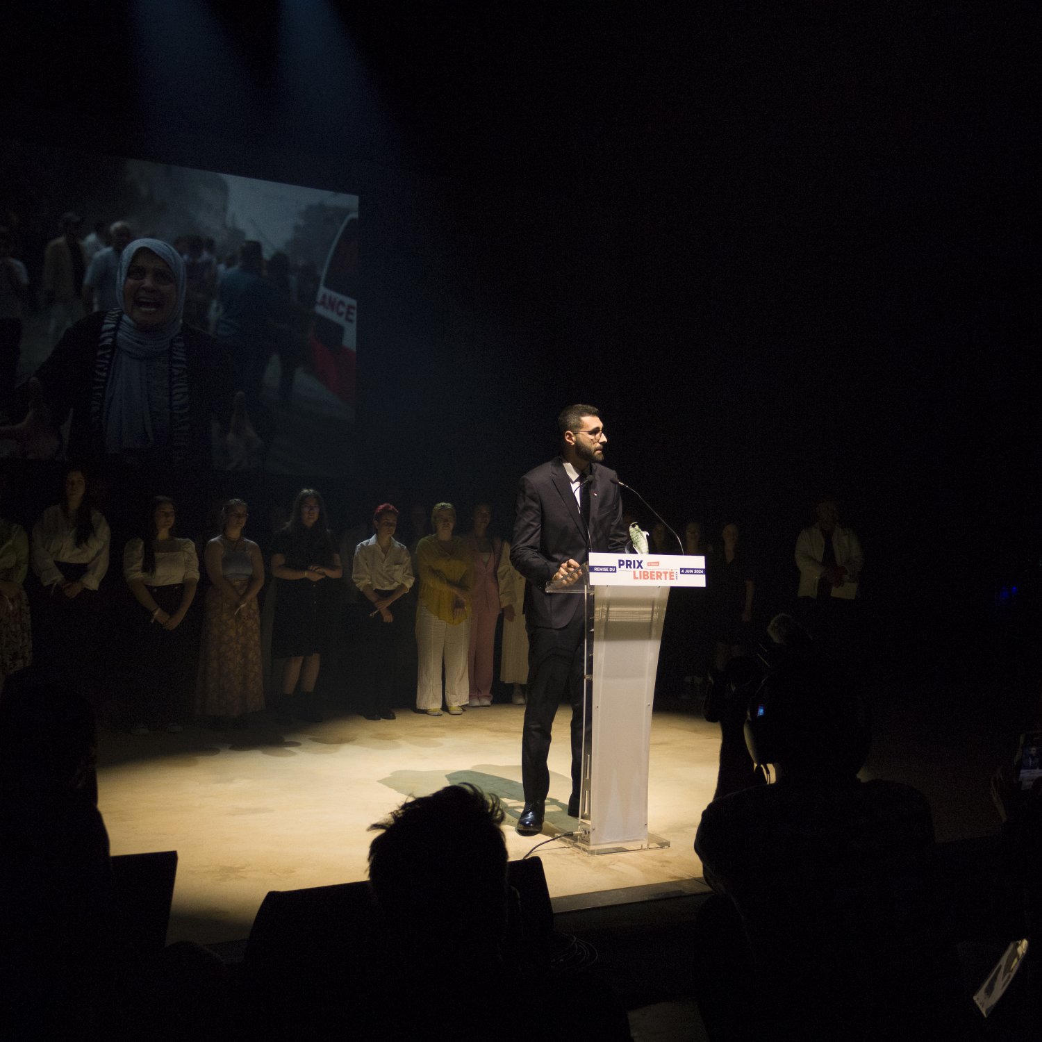 L'image montre un homme debout à un pupitre transparent, prenant la parole lors d'un événement. Il est habillé en costume et semble s'adresser à un public. En arrière-plan, on peut voir un écran projetant une vidéo ou une image, vraisemblablement liée à un sujet important, étant donné l'expression des personnes présentes. Au premier plan, on peut apercevoir des membres du public et des photographes, créant une atmosphère de concentration et d'engagement. L'éclairage met en évidence l'orateur, tandis que le reste de la scène est plus sombre.