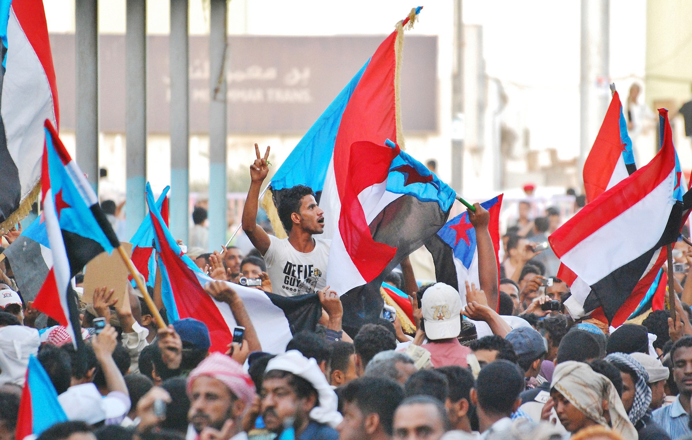 L'image montre une foule rassemblée, brandissant des drapeaux d'un pays ou d'une région, probablement en lien avec une démonstration ou une manifestation. Au centre, un homme se démarque en levant deux doigts en signe de victoire. La scène est animée, avec beaucoup de personnes exprimant leur enthousiasme ou leur engagement, ce qui indique une ambiance de protestation ou de célébration. Les couleurs des drapeaux sont vives, ce qui ajoute à l'énergie de la scène.