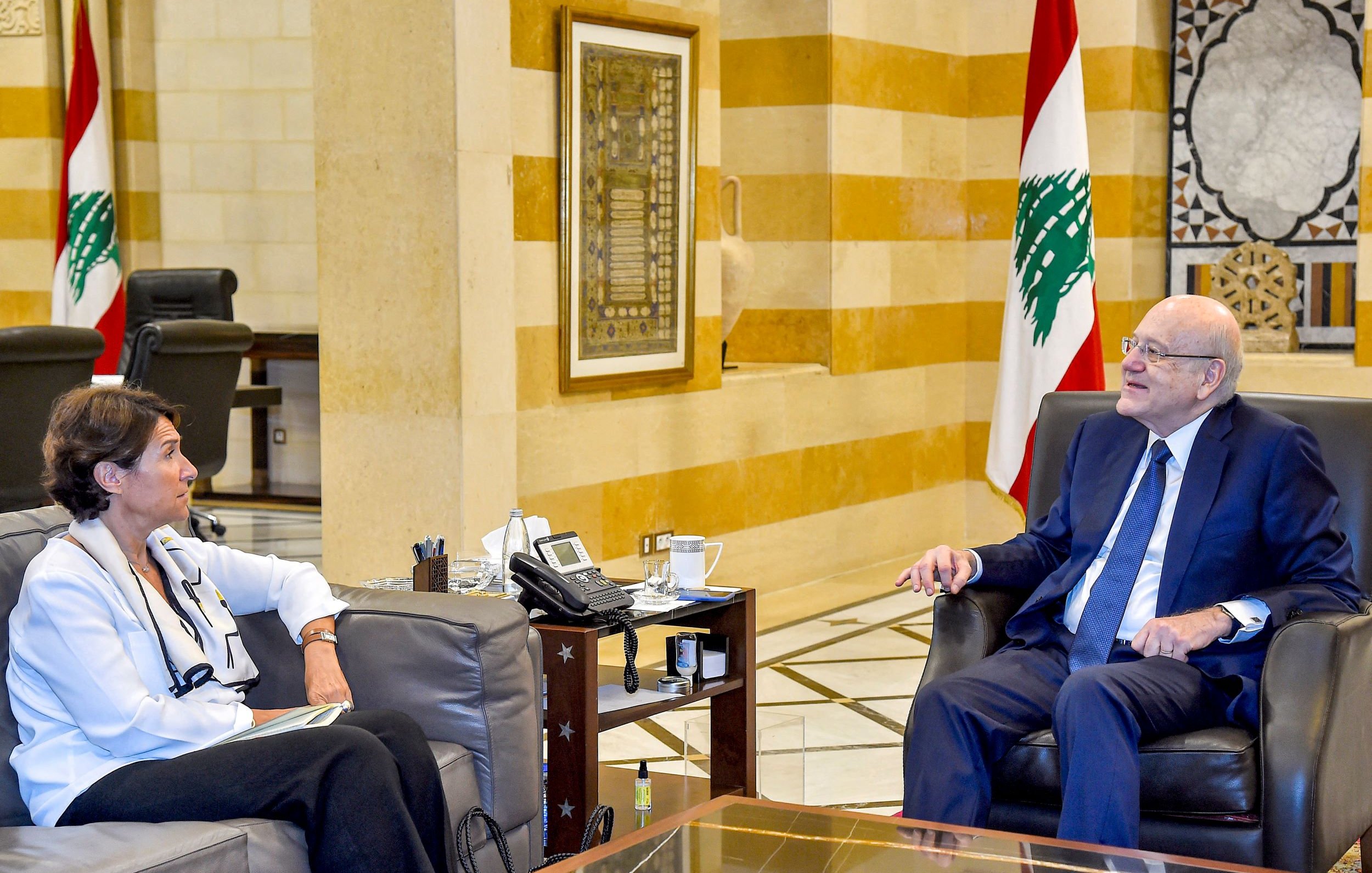 L'image montre une rencontre entre deux personnes assises dans un intérieur soigné, probablement dans un bureau ou une salle de réunion. L'un des individus est un homme en costume, tandis que l'autre est une femme habillée en chemise blanche. Ils semblent engagés dans une discussion, avec des expressions attentives. À l'arrière-plan, on peut apercevoir des drapeaux libanais et une décoration architecturale typique. La scène dégage une ambiance professionnelle et formelle.