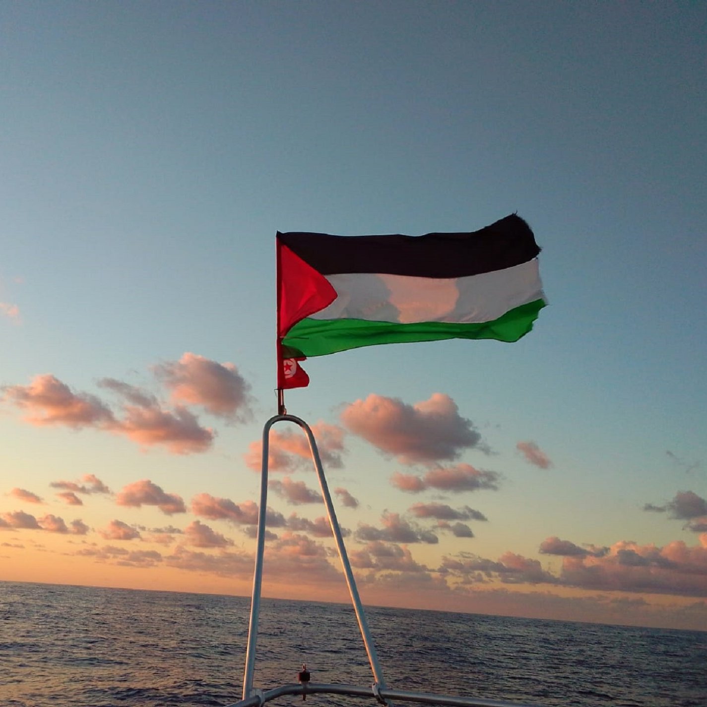 Drapeau palestinien flottant au-dessus de la mer, ciel aux teintes orangées et nuages.