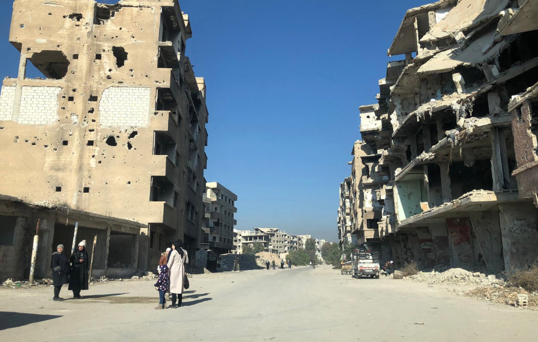 L'image montre une rue dévastée par des conflits, avec des bâtiments en ruines de part et d'autre. Les murs des bâtiments sont marqués par des impacts de balles et des débris sont éparpillés sur le sol. Au centre de la rue, quelques personnes marchent, tandis que des voitures abandonnées sont visibles. Le ciel est dégagé, ce qui contraste avec la destruction environnante.