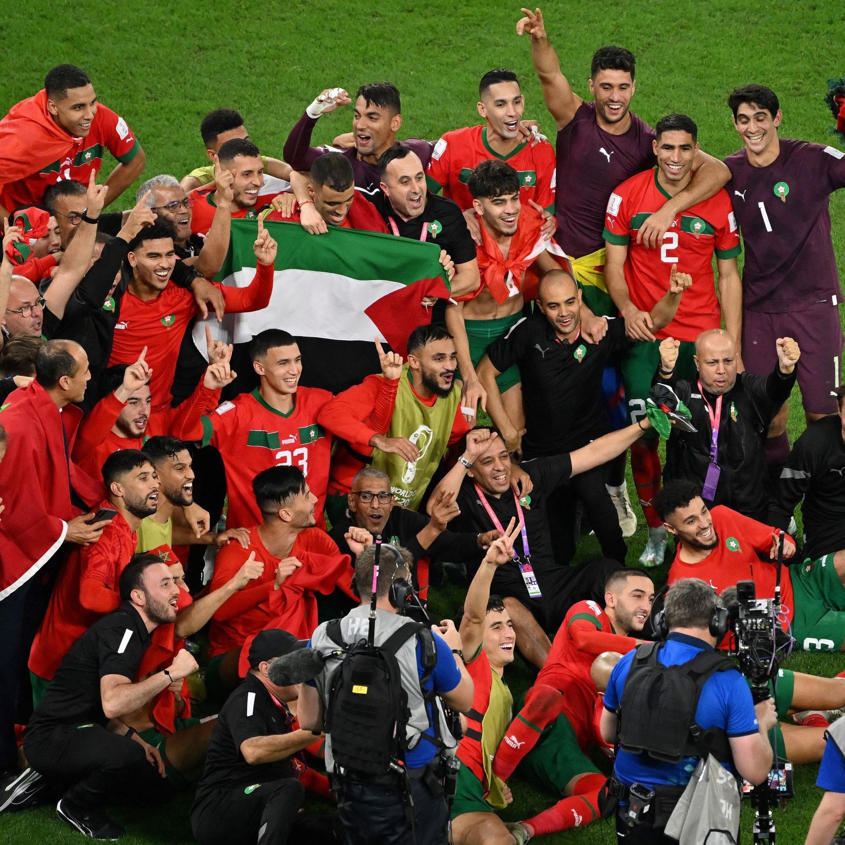 L'image montre une équipe de football, probablement celle du Maroc, célébrant une victoire. Les joueurs sont rassemblés, certains brandissant un drapeau marocain et d'autres levant les bras en signe de joie. Ils sont entourés de membres du staff et de caméras, capturant ce moment de fête. L'atmosphère est festive et dynamique, remplie d'excitation et de camaraderie.