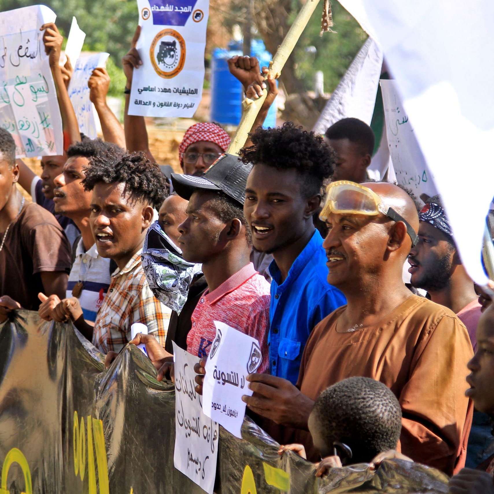 در این تصویر گروهی از مردم در حال برگزاری تجمع یا تظاهرات هستند. آن‌ها پلاکاردهایی در دست دارند که روی آن‌ها نوشته‌هایی به زبان عربی وجود دارد. به نظر می‌رسد که این جمعیت شامل مردان جوان و بزرگسال است که برخی از آن‌ها لباس‌های سنتی و برخی لباس‌های عادی پوشیده‌اند. فضای تصویر نشان‌دهنده شور و هیجان جمعیت است و احتمالا این تظاهرات به یک مسئله اجتماعی یا سیاسی مرتبط است.