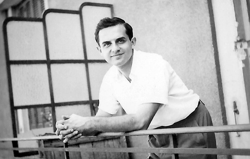 L'image montre un jeune homme posant avec un sourire amical. Il est vêtu d'une chemise blanche et de pantalons clairs, se tenant sur un balcon avec des barres métalliques. L'arrière-plan présente une architecture simple, avec des fenêtres et des volets. L'ambiance est décontractée et ensoleillée, suggérant un moment de détente.