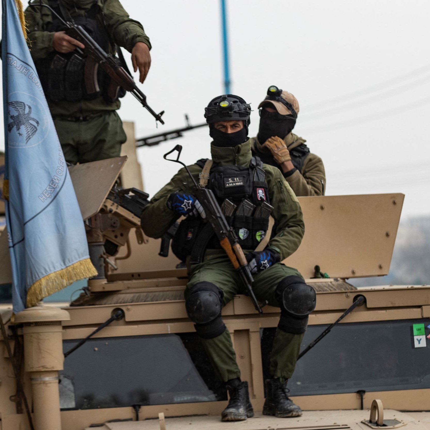 L'image montre des soldats en uniforme, armés et équipés de casques, se trouvant sur un véhicule militaire. L'un des soldats est assis sur le toit du véhicule, tenant une arme, tandis que d'autres soldats sont debout à proximité. On peut également voir un drapeau, probablement lié à une mission de maintien de la paix. L'environnement semble urbain, avec des éléments de la ville en arrière-plan.