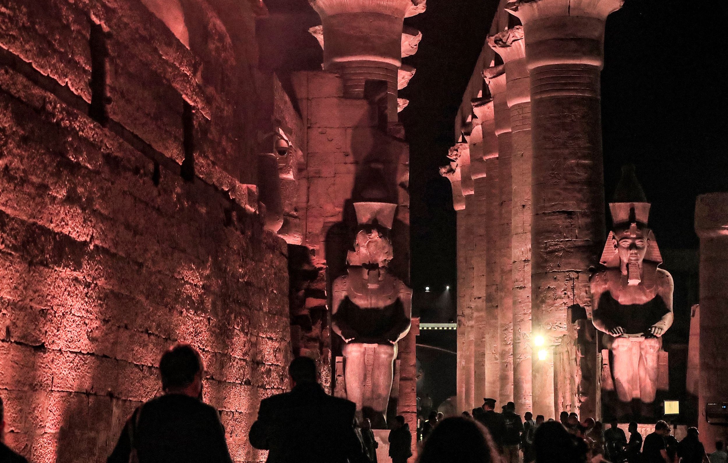 L'image présente un site historique, probablement des ruines égyptiennes, éclairées par des lumières tamisées, ce qui crée une atmosphère mystérieuse. On peut voir de grandes statues de pharaons ou de divinités, flanquant une structure impressionnante faite de colonnes. Les visiteurs se déplacent à travers le site, ajoutant une dimension humaine à cette scène ancienne, où l'architecture et les sculptures témoignent de la grandeur de la civilisation égyptienne. Les ombres et la lumière jouent un rôle important, accentuant les détails des sculptures et des murs.