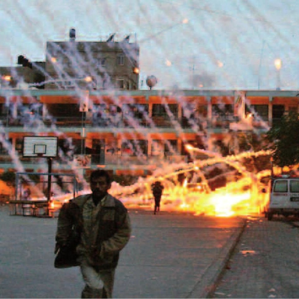 L'image montre une scène de chaos avec des explosions et des traînées de lumière dans le ciel. Des personnes semblent fuir dans une zone qui ressemble à une cour d'école, tandis qu'un bâtiment en arrière-plan est visible. L'environnement semble marqué par une tension intense, avec des traces d'incendie et des éclats lumineux qui suggèrent une situation dangereuse. L'atmosphère est sombre, renforçant le sentiment d'urgence et de désarroi.