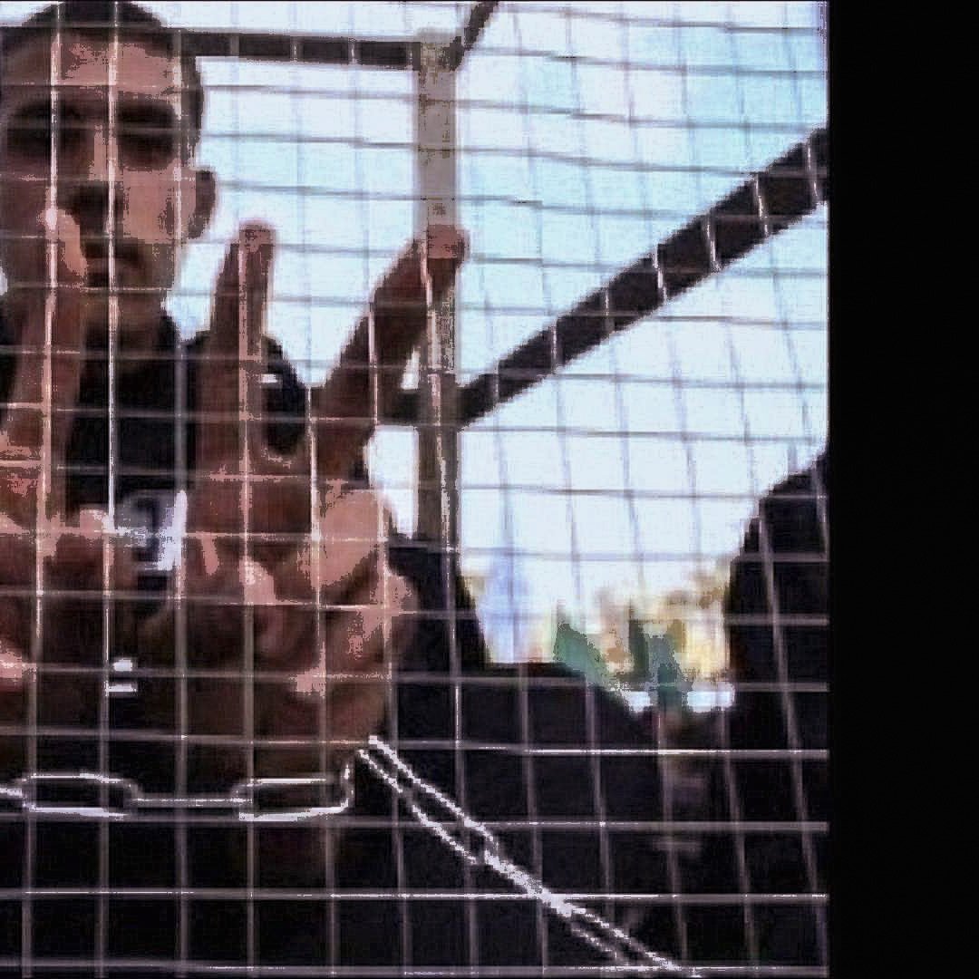 الصورة تظهر شابًا يقف خلف شبكة أو قفص، حيث يرفع يديه مكونًا علامة أو عددًا معينًا بإشارات أصابعه. يبدو أنه محاط بمساحة ضيقة، مما يعطي إيحاءً بالاحتجاز أو العزلة. تعكس تعابير وجهه أو وضع يديه مشاعر معينة، مثل المقاومة أو الإصرار. الخلفية غير واضحة تمامًا، مما يركز الانتباه على الشاب وإشارته.