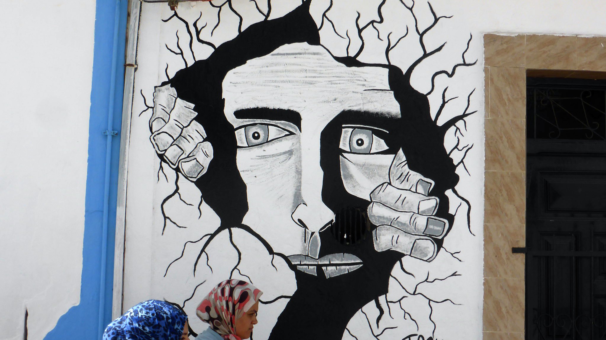 L'image montre une fresque murale en noir et blanc représentant un visage humain au milieu d'une apparente fissure dans le mur. Le visage a des traits marqués et semble exprimer une émotion intense. Des mains émergent des fissures, comme si elles tentaient de sortir. Au premier plan, deux femmes en hijab marchent, apportant une touche de couleur et de vie à la scène. Le tout crée un contraste entre l'art mural et la réalité environnante.