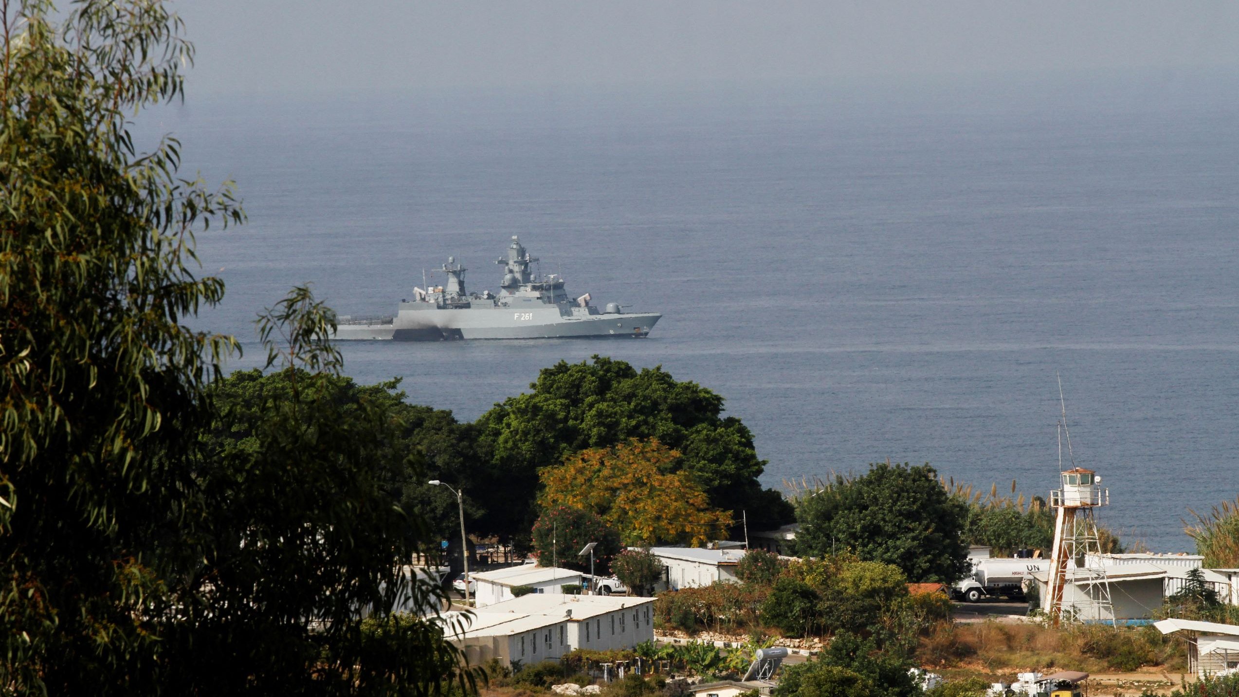در تصویر، یک کشتی جنگی در نزدیکی ساحل دیده می‌شود که در دریا شناور است. این کشتی دارای طراحی مدرن و تجهیزات نظامی است. در پیش‌زمینه، تعدادی درخت و ساختمان‌های کوچک دیده می‌شود، و پس‌زمینه نمایی از دریا و آسمان روشن دارد. تضاد بین رنگ‌های سبز درختان و آبی دریا جلوه‌ای زیبا به تصویر بخشیده است.