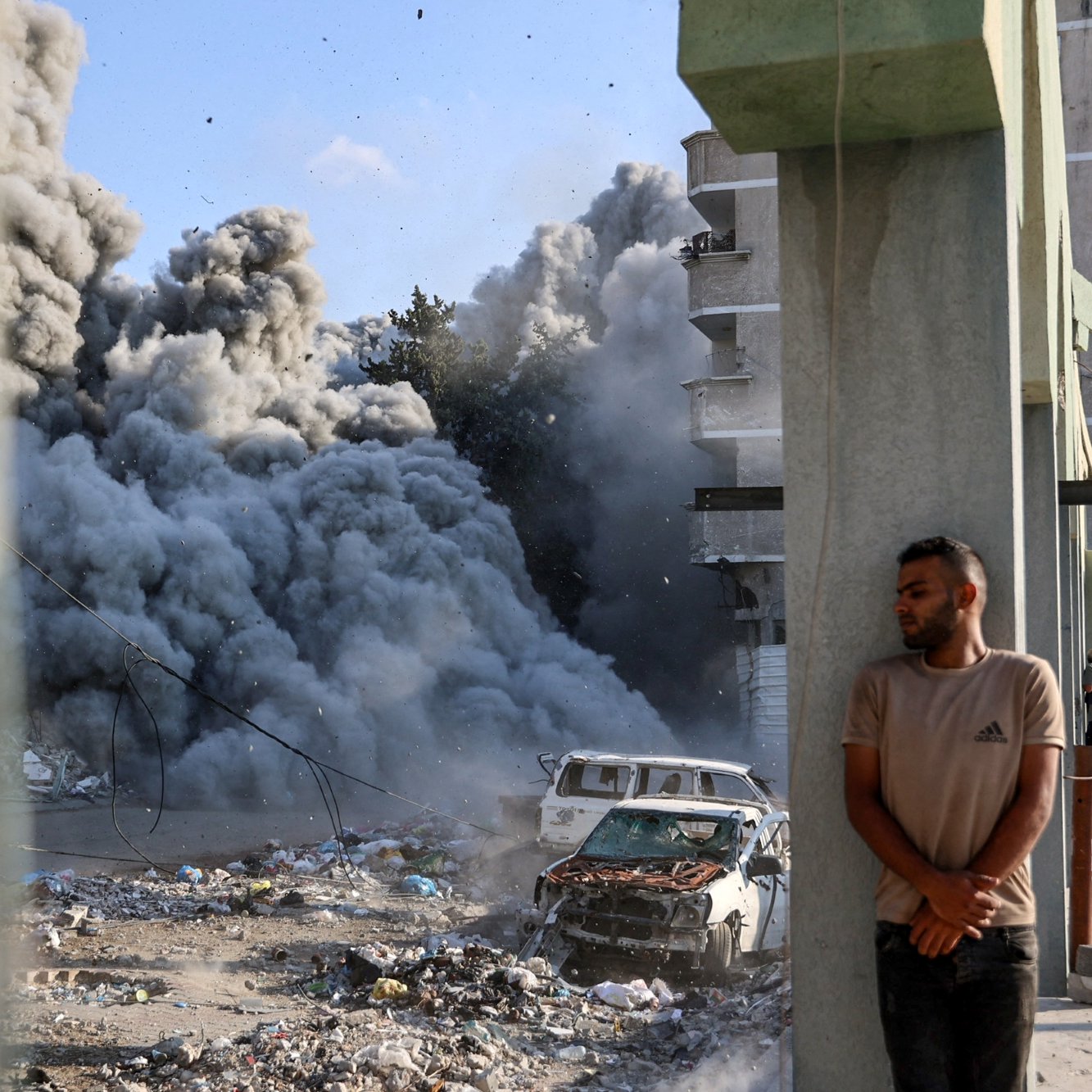 Destruction urbaine avec de la fumée, un homme adossé à un pilier près d'une voiture endommagée.