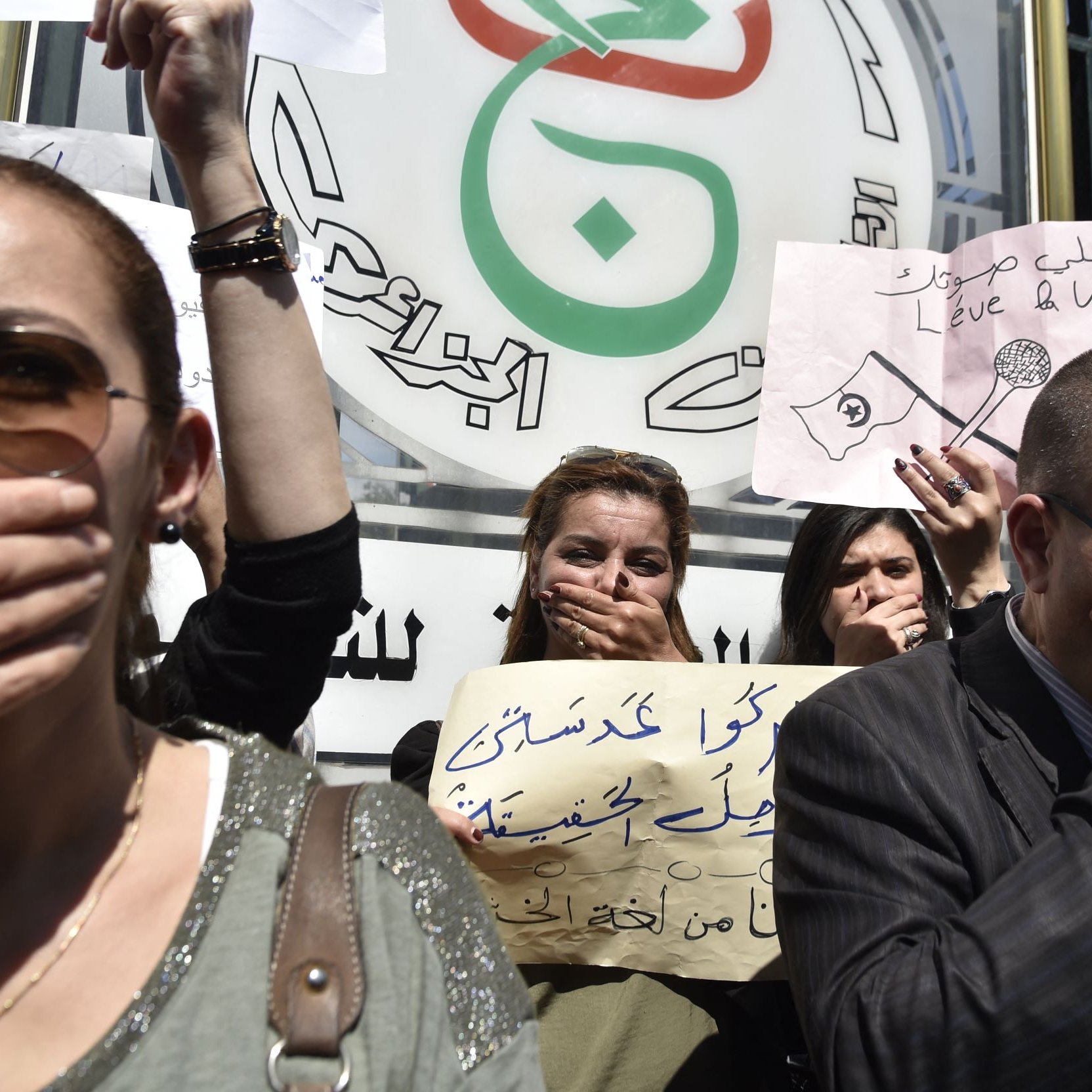 L'image montre une manifestation avec des personnes tenant des pancartes. Elles portent toutes la main sur la bouche, symbole de protestation ou de silence forcé. On peut voir des messages écrits sur les pancartes, qui semblent exprimer des revendications ou des critiques. Les manifestants sont sérieux et déterminés, reflétant un fort engagement pour leur cause. En arrière-plan, il y a un logo ou un symbole en rapport avec le mouvement.