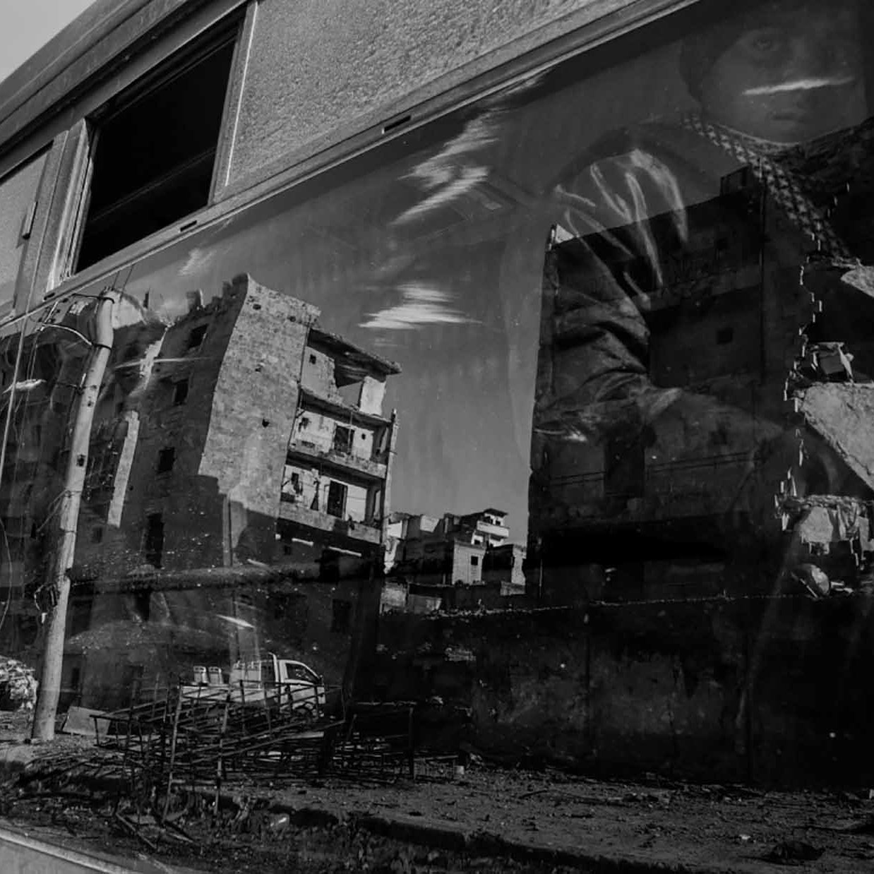 الصورة تظهر منظرًا حضريًا بواسطة زجاج نافذة حافلة. تعكس الصورة مجموعة من المباني المتضررة، مما يدل على دمار أو تدهور في المنطقة. يظهر أيضاً شخص يجلس بجانب النافذة، وملامح وجهه غير واضحة بسبب الانعكاس. الألوان في الصورة تتسم باللونين الأبيض والأسود، مما يضفي طابعاً درامياً وأجواءً حزينة على المشهد.