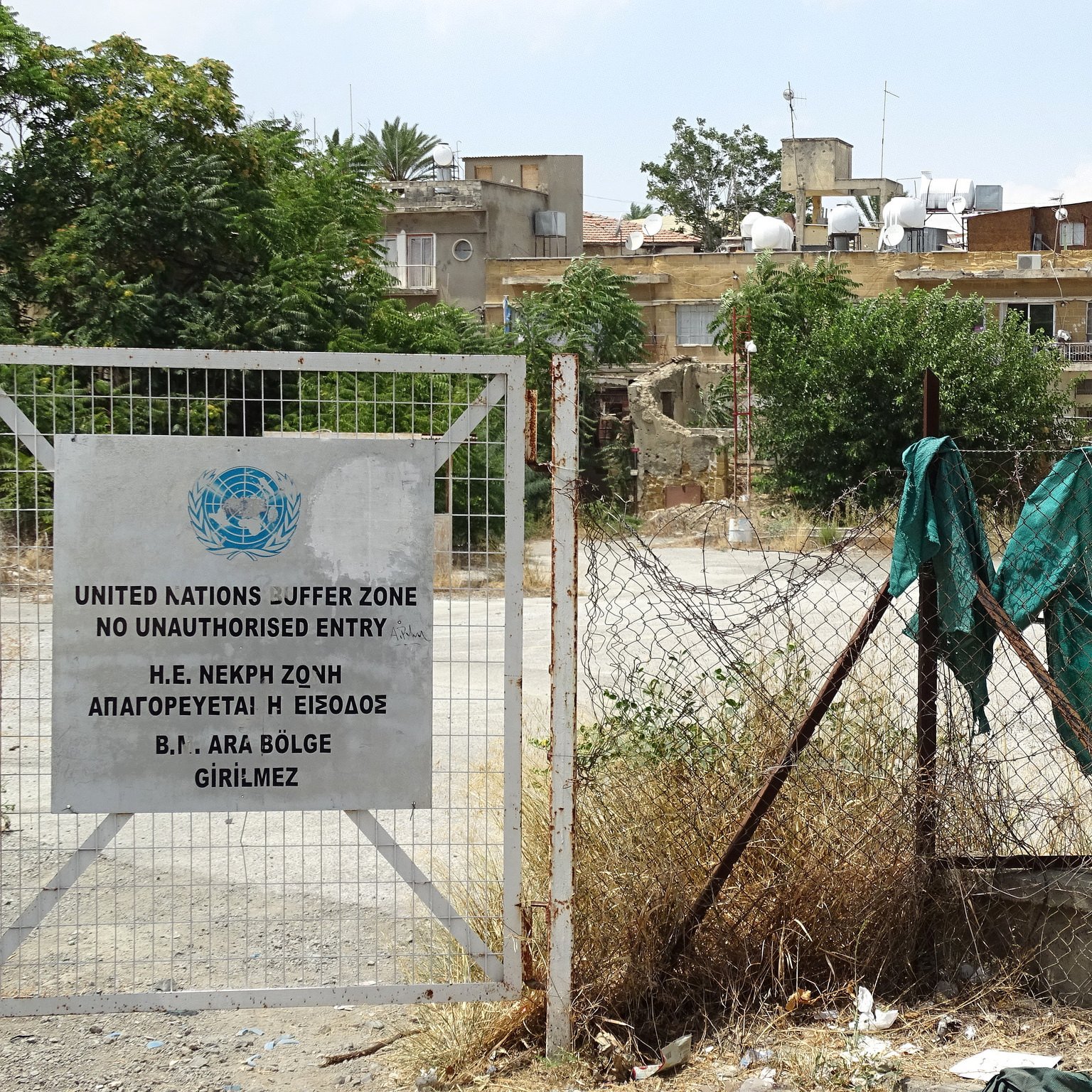 L'image montre une zone entourée d'une clôture. À gauche, il y a un panneau de la zone tampon des Nations Unies, indiquant qu'il s'agit d'un espace où l'entrée est interdite sans autorisation. À l'arrière-plan, on peut voir des bâtiments avec des antennes sur le toit, ainsi que de la végétation désordonnée au sol, créant une atmosphère de négligence. Le tout semble évoquer une région où l'accès est strictement contrôlé, probablement en raison de tensions historiques.